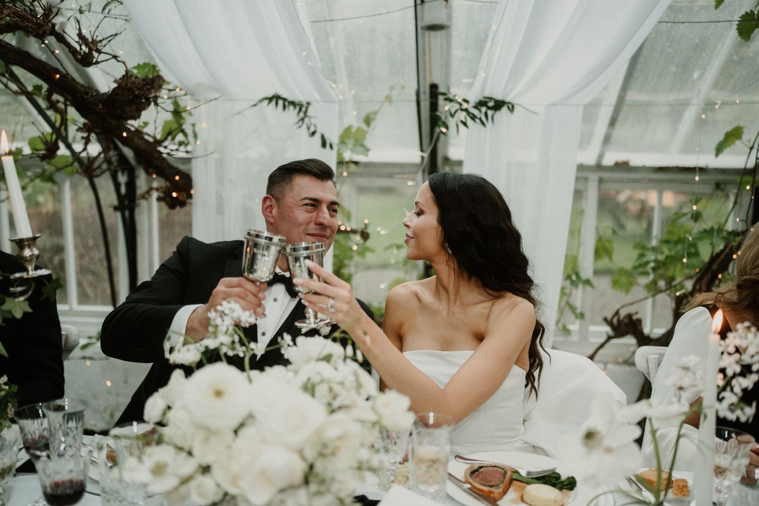 Couple toasts at their wedding reception surrounded by white floral decor and draped fabric.