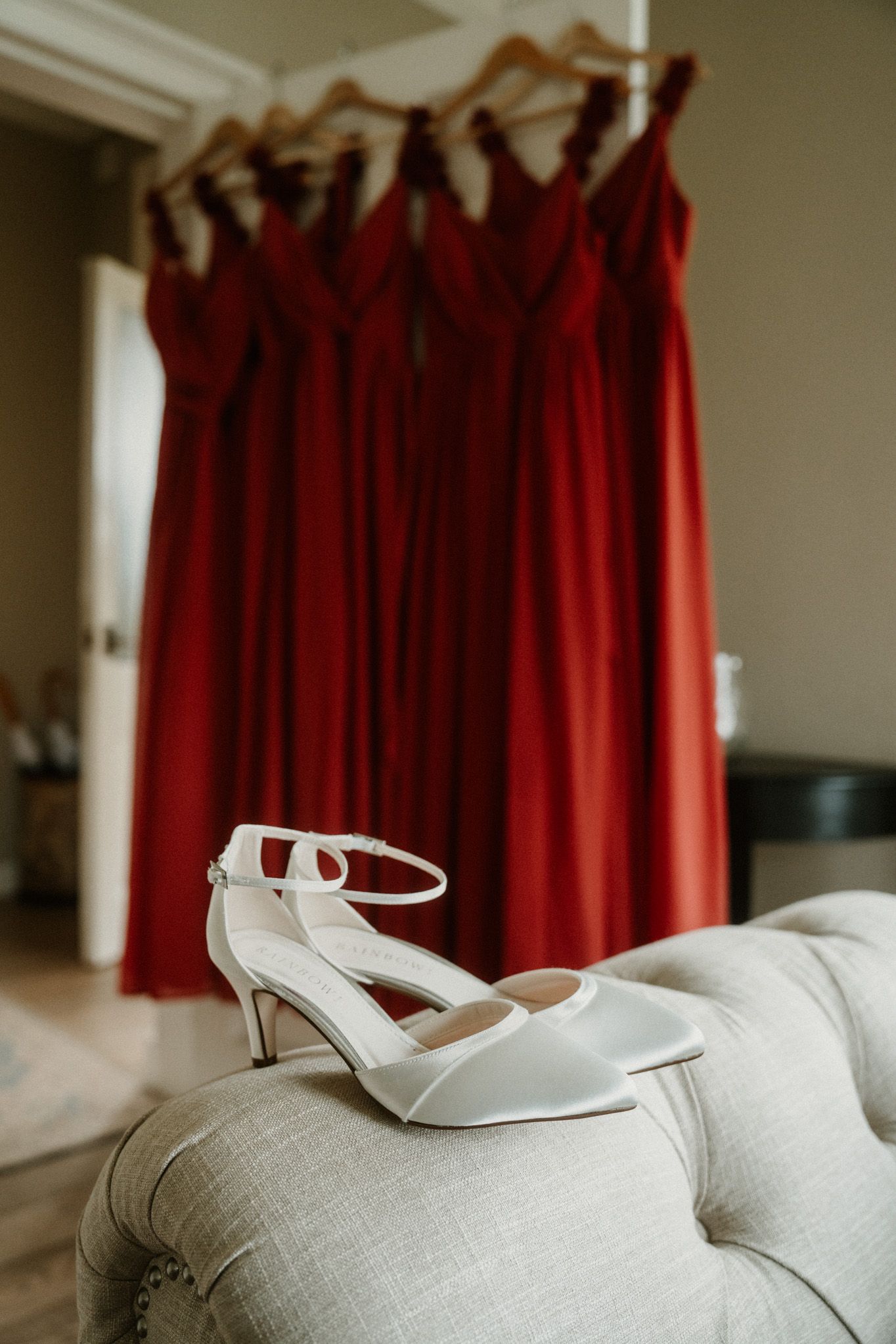 White bridal shoes displayed against red bridesmaid dresses hanging in preparation room.