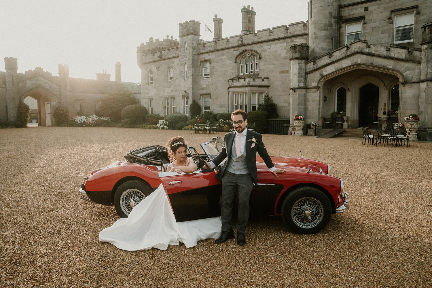 Classic red vintage sports car parked in front of a historic stone castle during a wedding celebration.