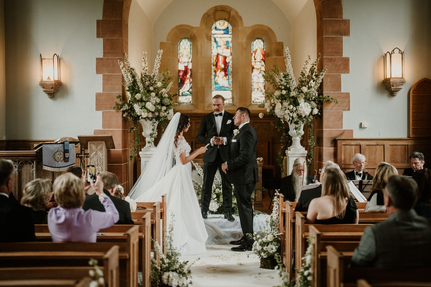 Guests watch as newlyweds walk down the aisle in a church decorated with white floral arrangements.