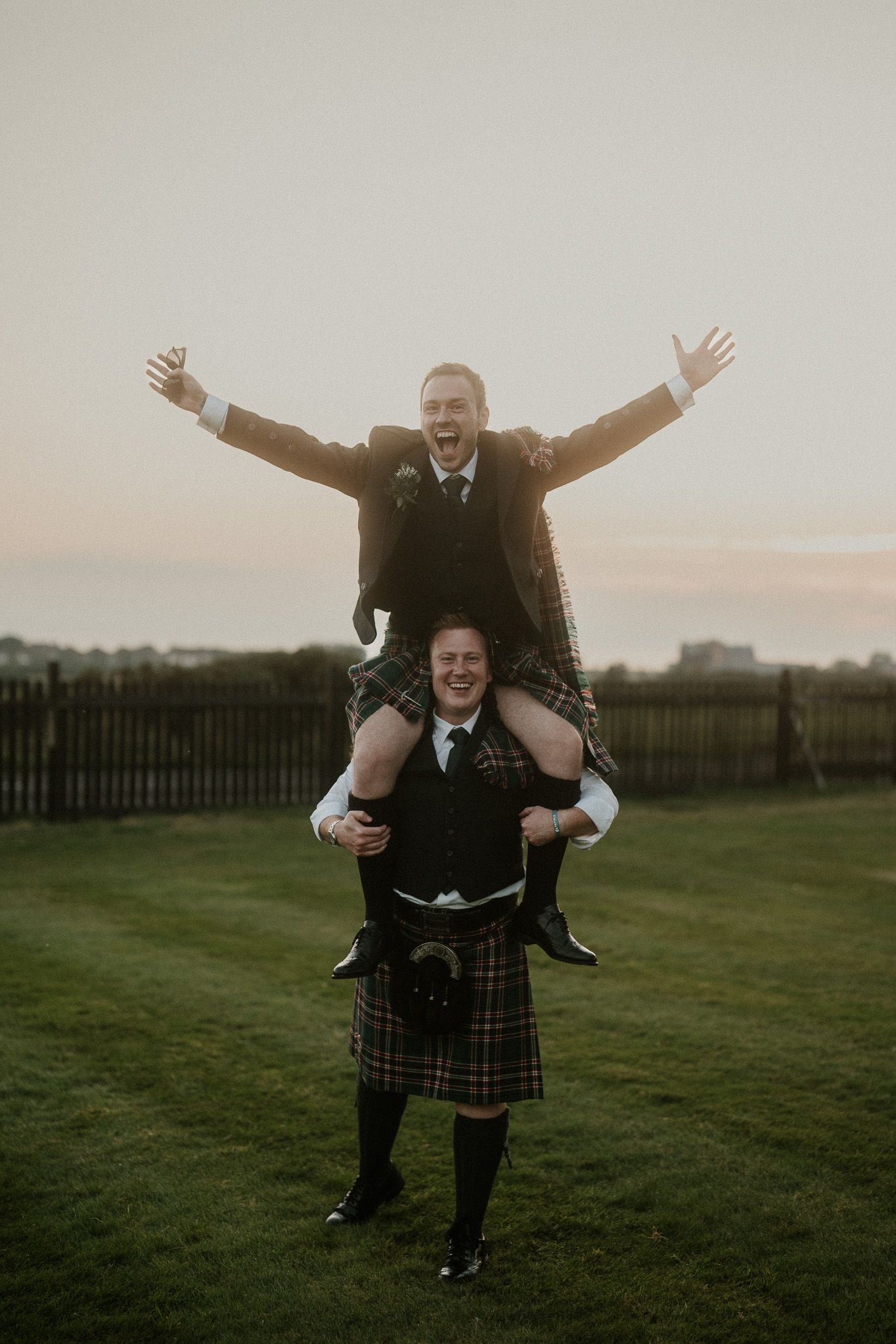 Two people in kilts celebrate joyfully with a piggyback ride on grassy grounds at sunset.
