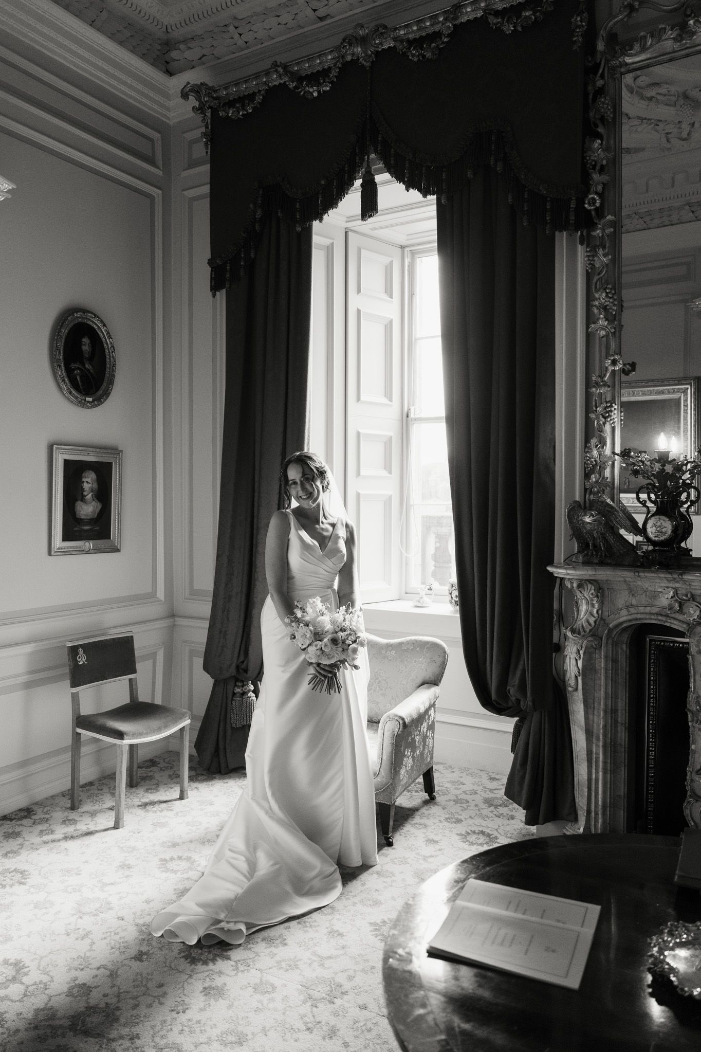 Black and white photo of a bride in a flowing gown holding a bouquet beside tall draped windows in an elegant room.