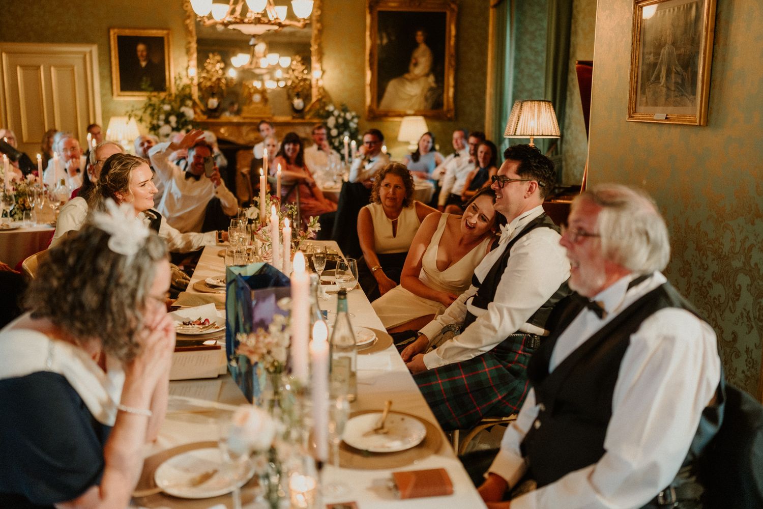 Wedding guests in formal attire laugh and enjoy a candlelit banquet at long tables in an elegant, portrait-lined dining room.