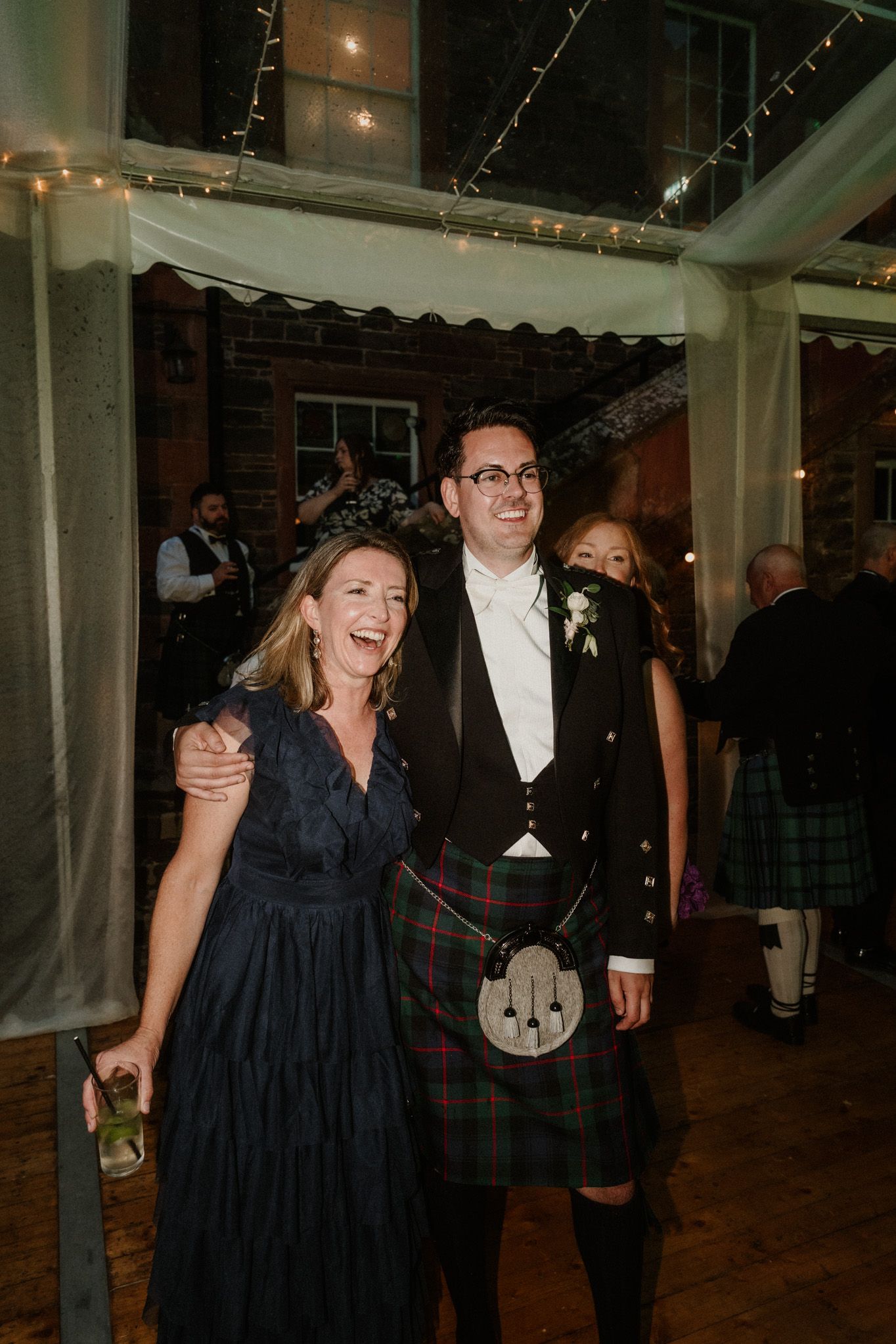 Laughing woman in a navy dress embraces a man in a kilt and tartan at a festive wedding reception under a string-lit tent.