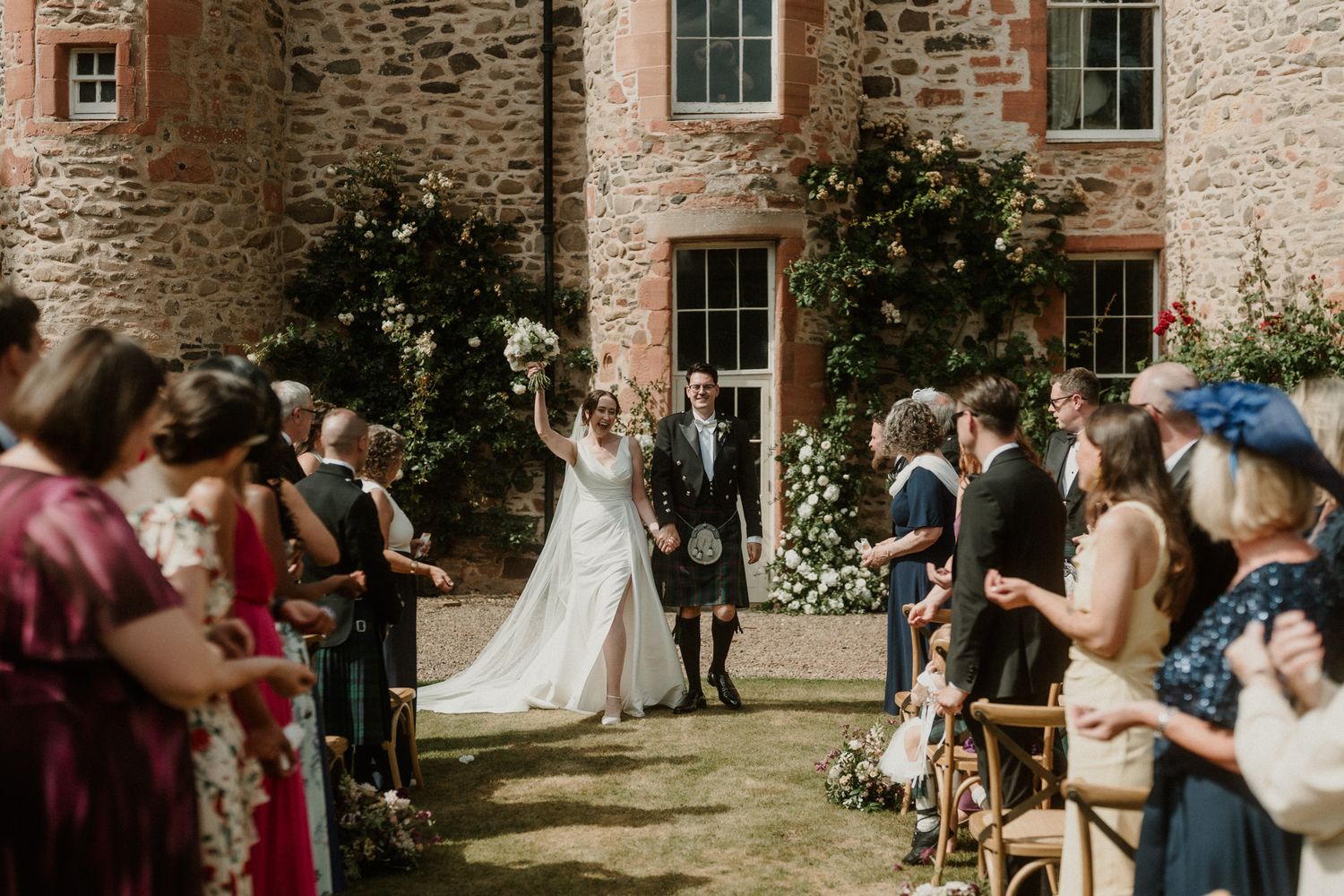 Bride in a white gown raises her bouquet while walking down an outdoor aisle with her groom, as guests cheer at a stone castle wedding.