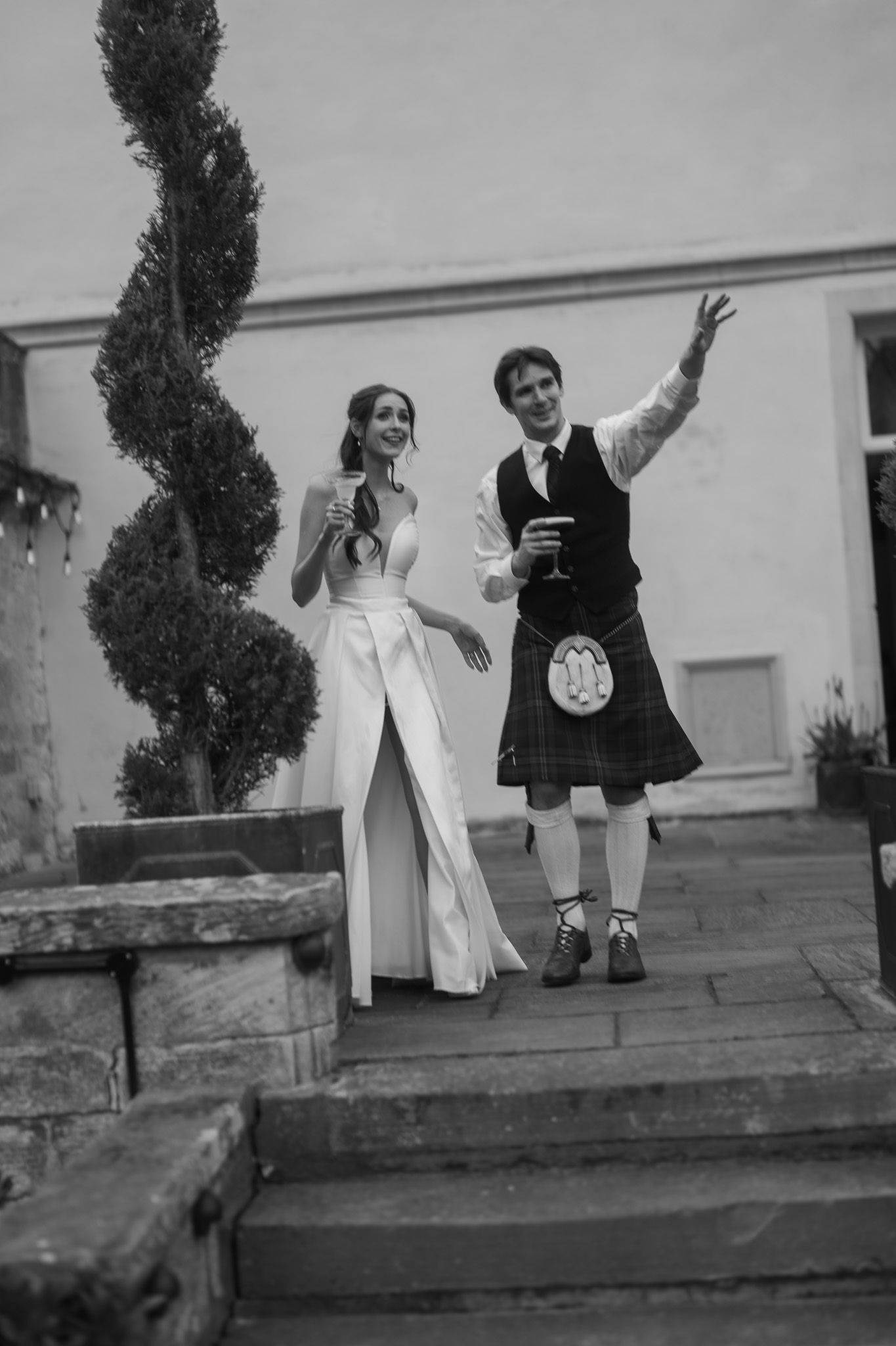 Black and white photo of a bride in white gown and groom in Scottish kilt raising glasses on stone steps.