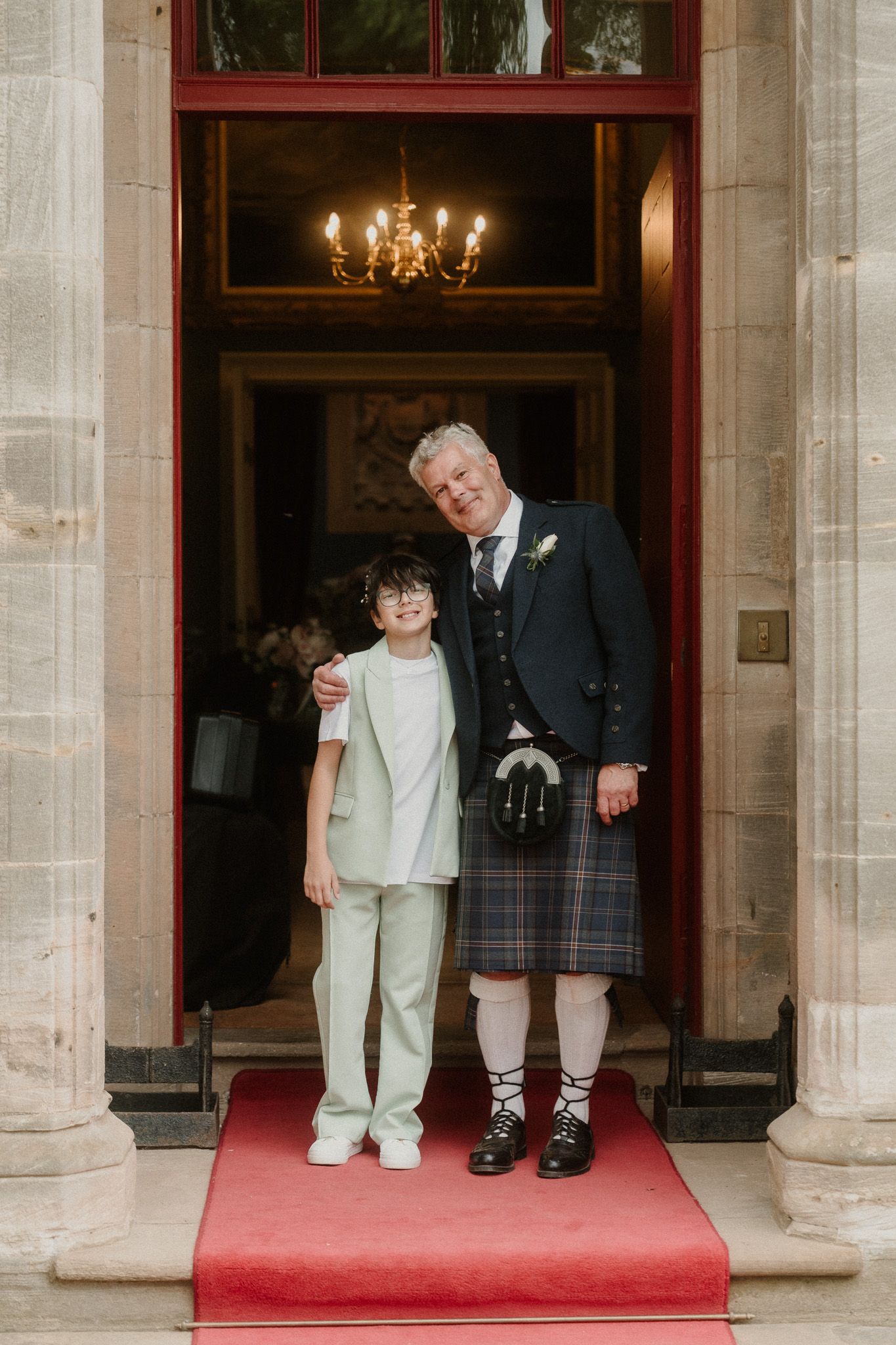 Man in Scottish kilt and boy in sage suit pose together on red carpet steps at a formal venue.