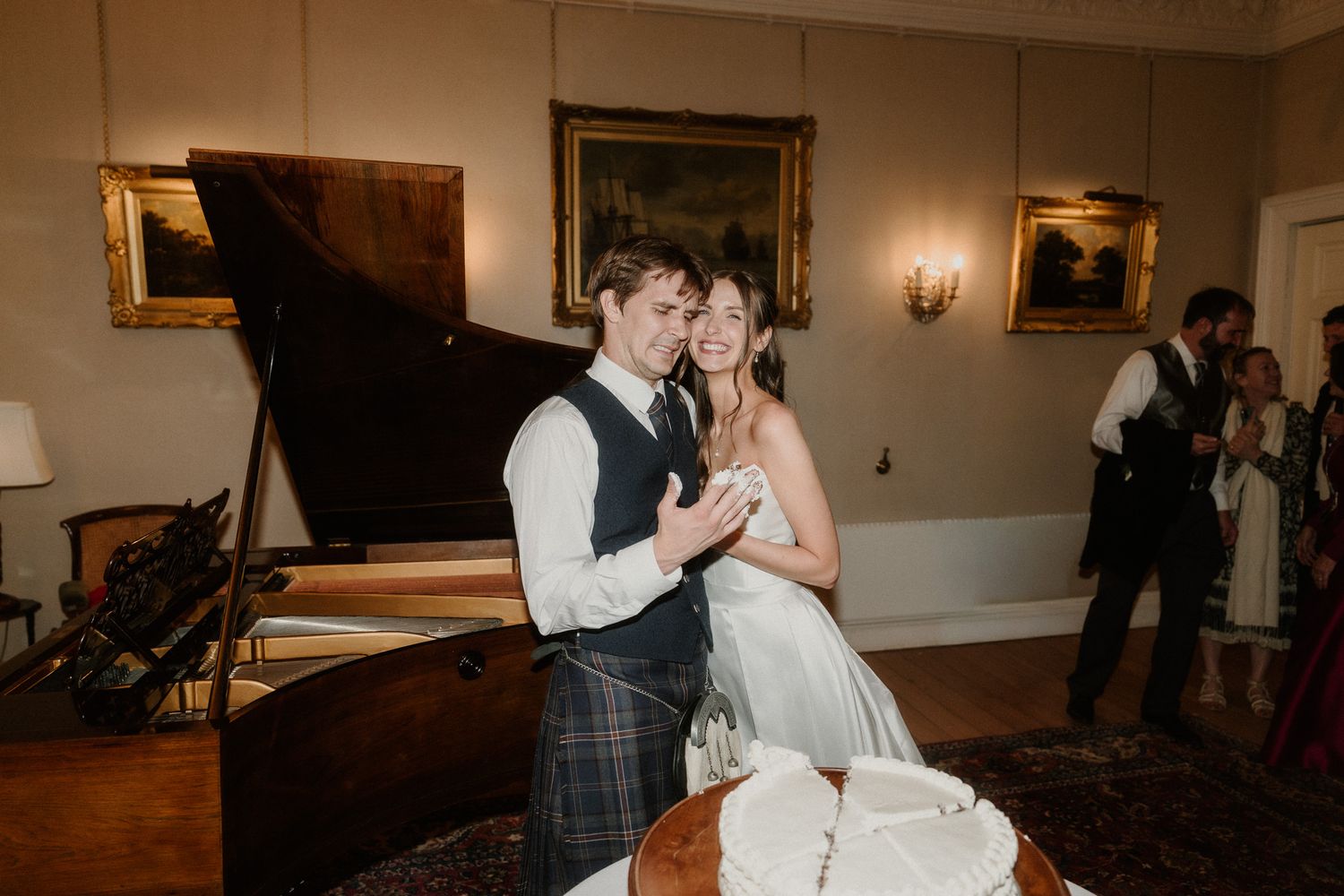 Bride and groom smile together near a grand piano and wedding cake at an elegant Scottish wedding reception.