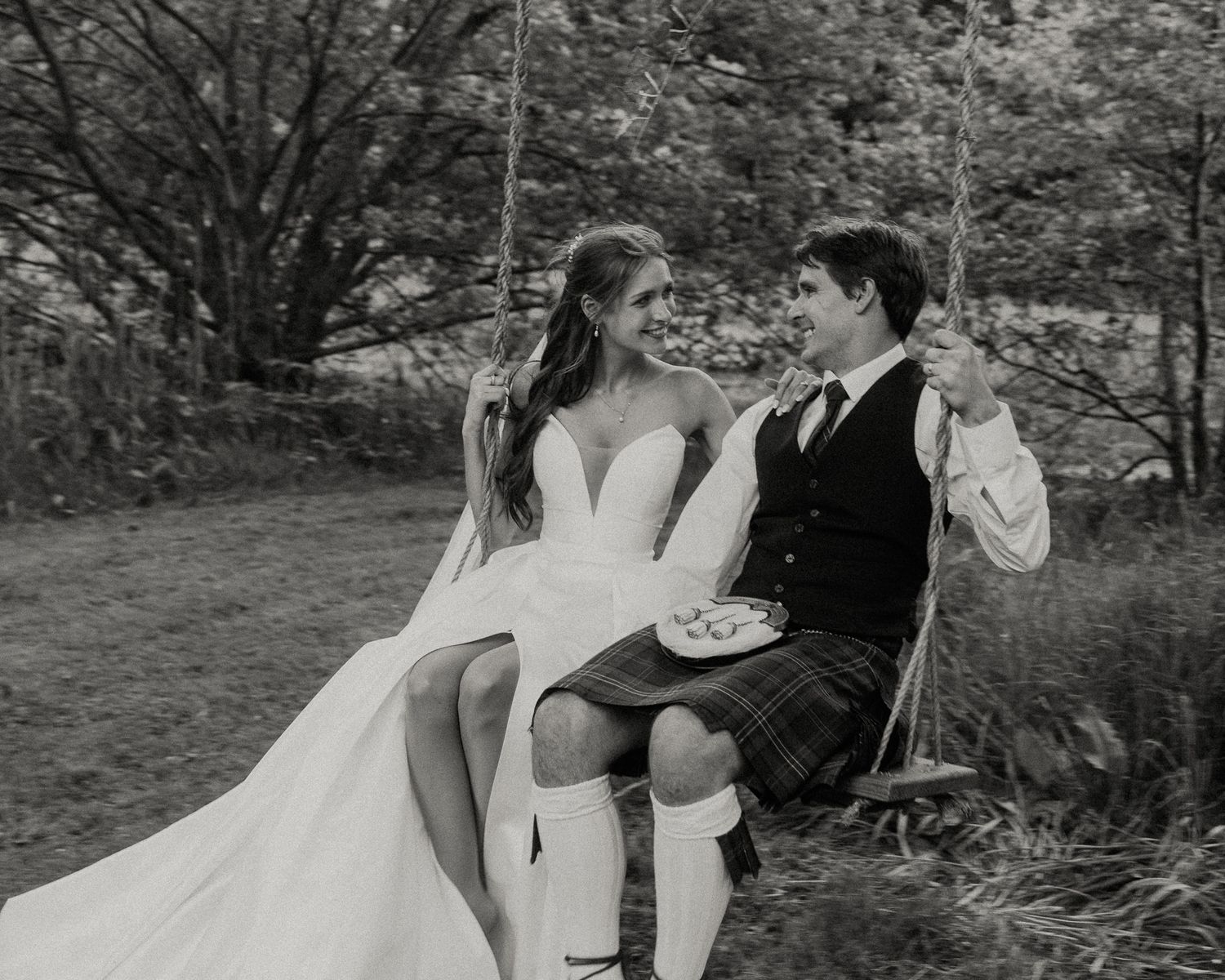 Black and white photo of bride and groom sitting on a garden swing, groom wearing a kilt, sharing a romantic moment.