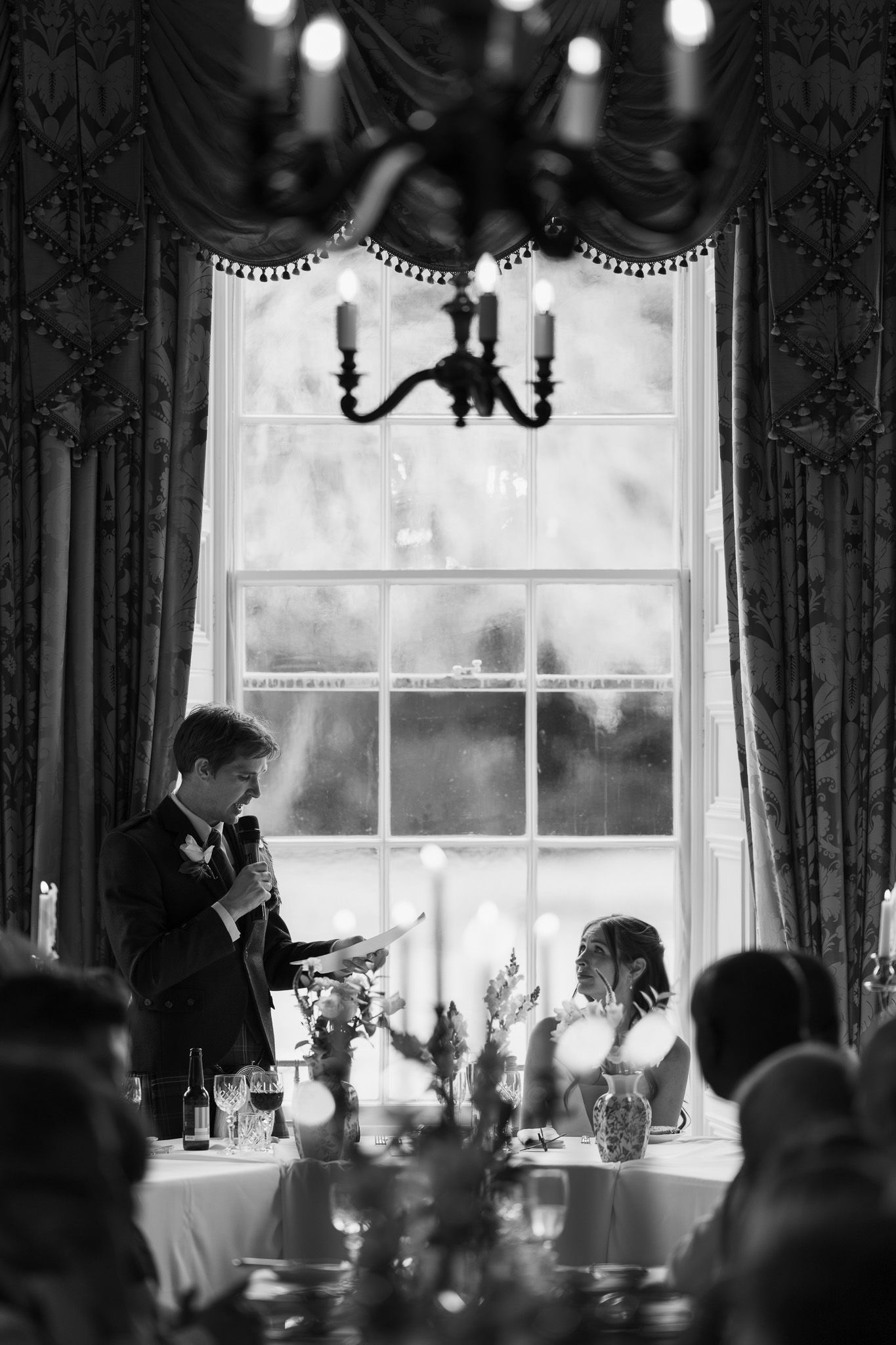 Black and white wedding speech photo with groom reading notes by elegant window with chandelier above.