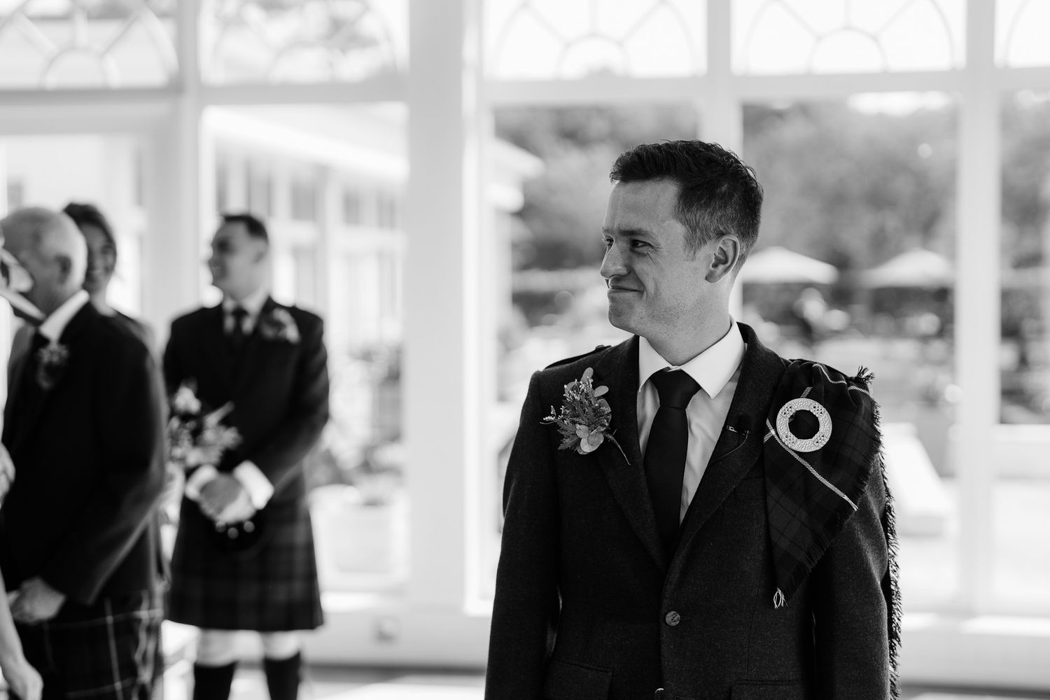 Wedding attendees in formal attire and kilts gather in a sunlit room.