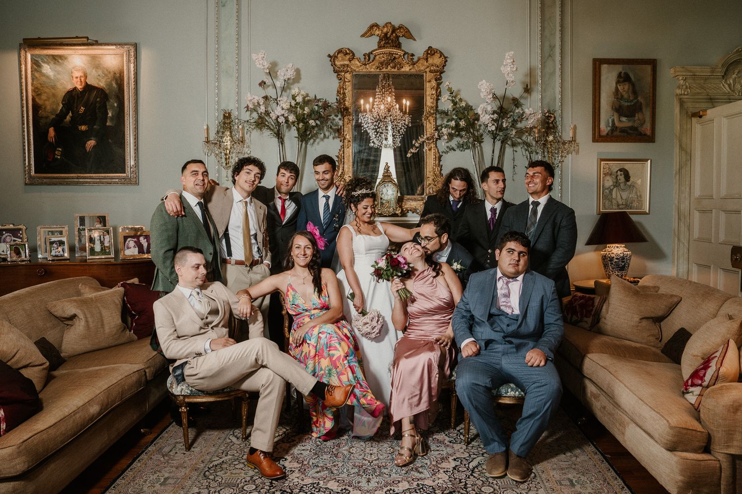 Wedding party members seated on vintage couches in formal drawing room for group photos.