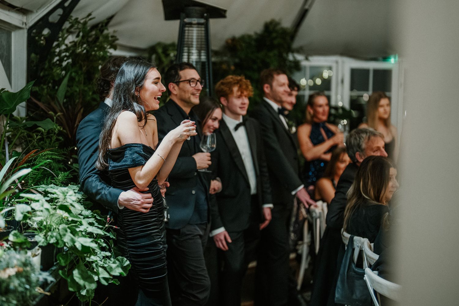 Formal wedding reception guests gathered in a dimly lit venue with greenery and elegant decor.
