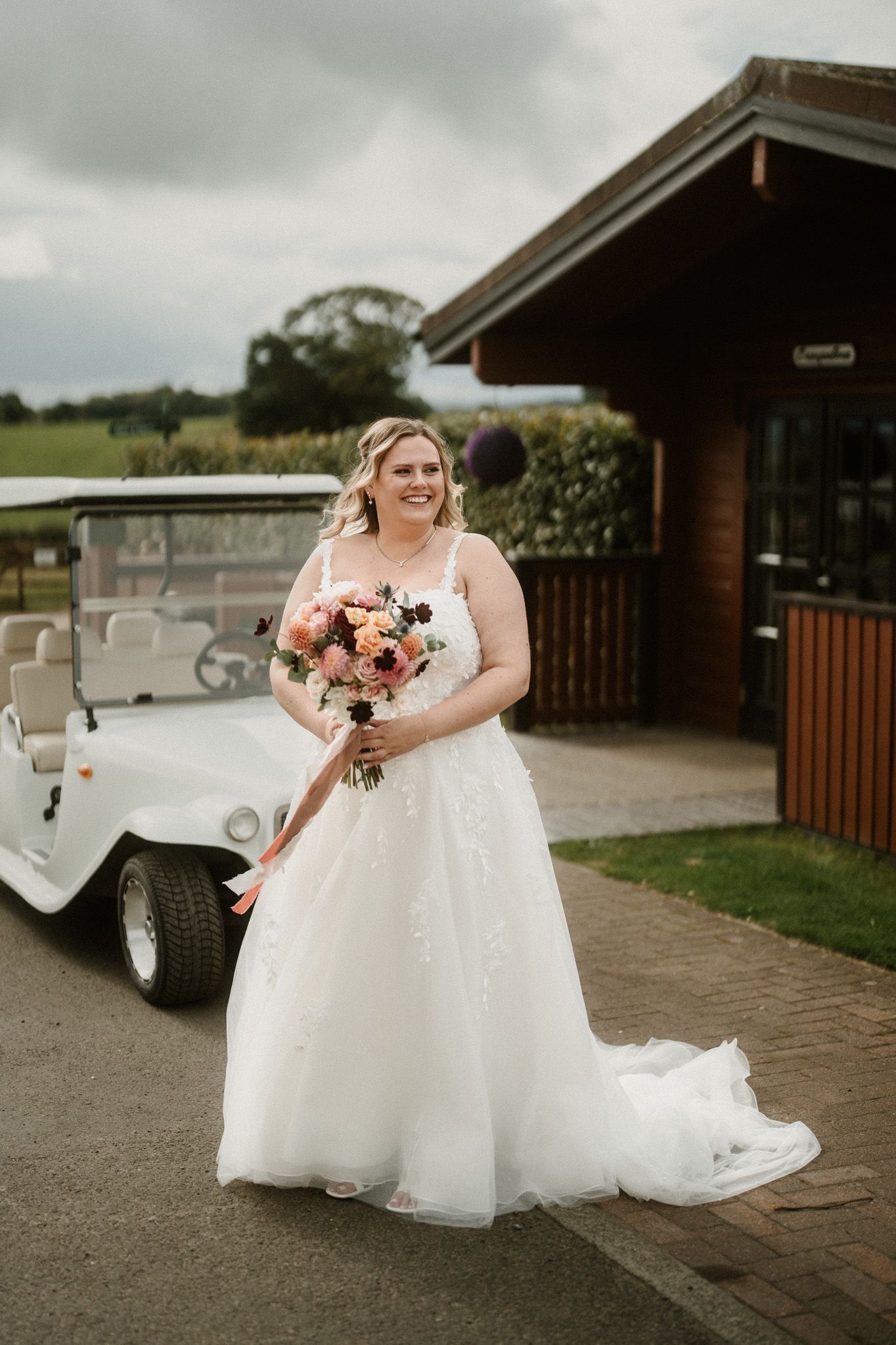 White wedding dress and bouquet photographed outside rustic venue with vintage car.