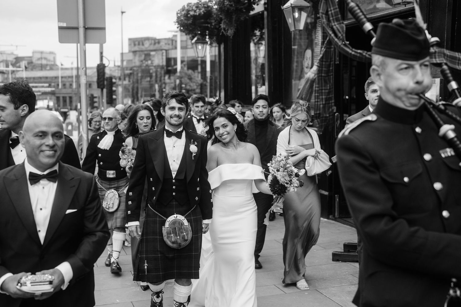 A bagpiper leads a wedding procession down a city street in traditional Scottish attire.