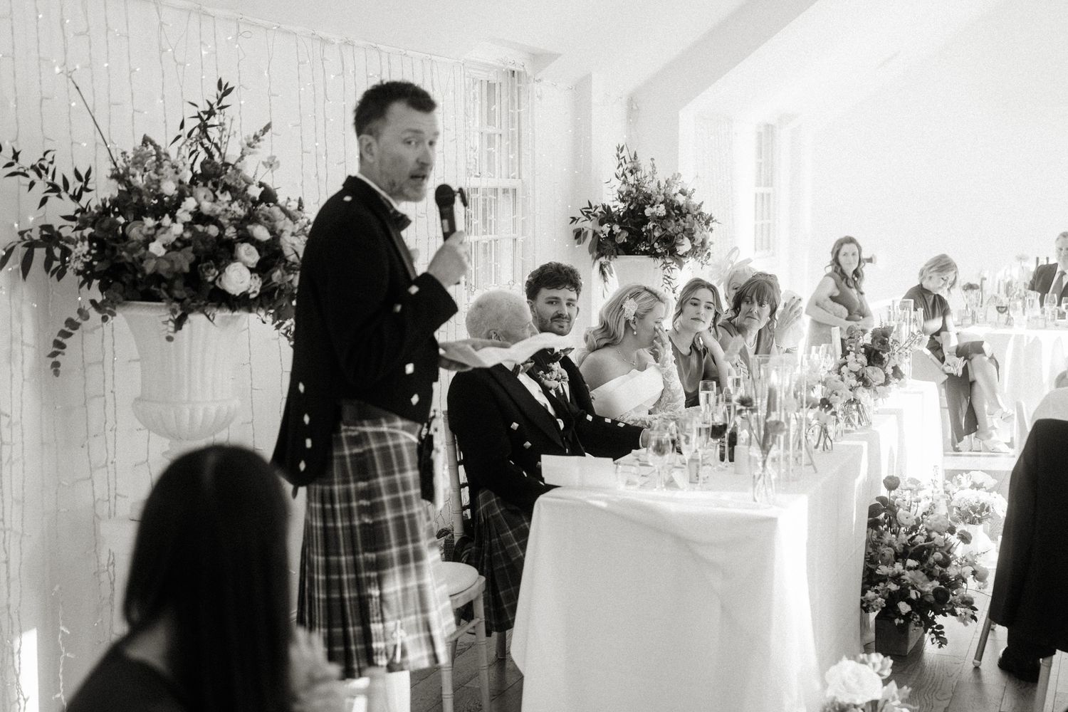 Wedding guests seated at decorated tables in a bright reception hall with floral arrangements.