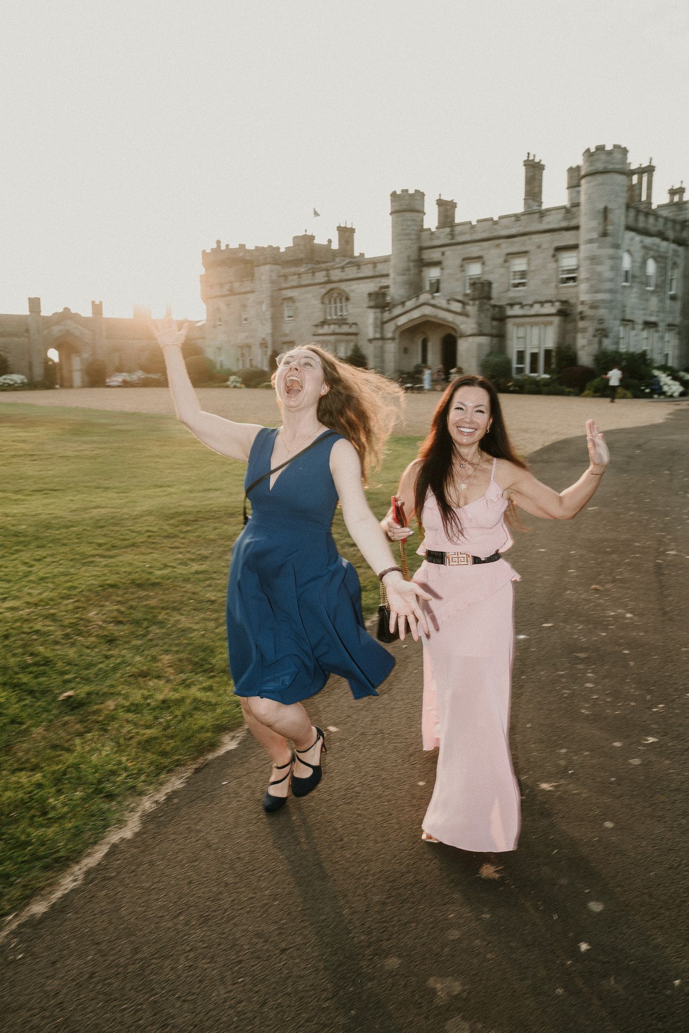 Two people dance joyfully on the lawn in front of a historic castle at sunset.