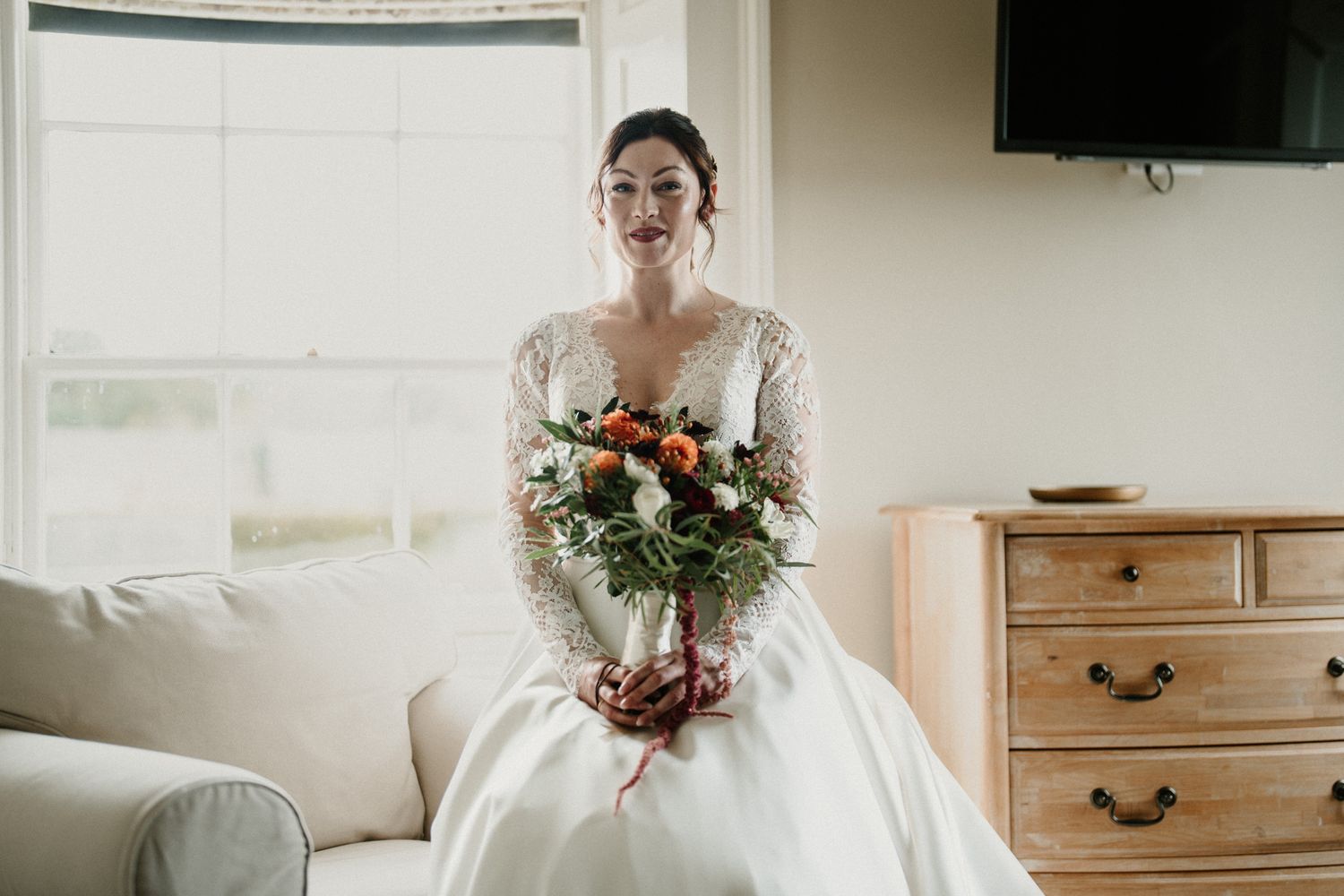 A wedding dress with lace details and full skirt photographed in a bright room with wooden furniture.