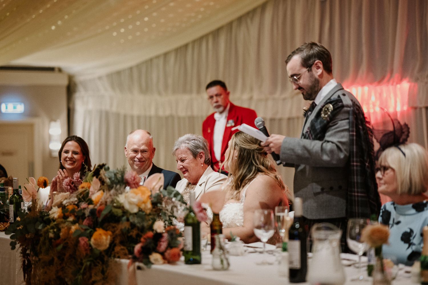 Wedding reception speech at head table with elegant floral centerpieces and draped ceiling decor.