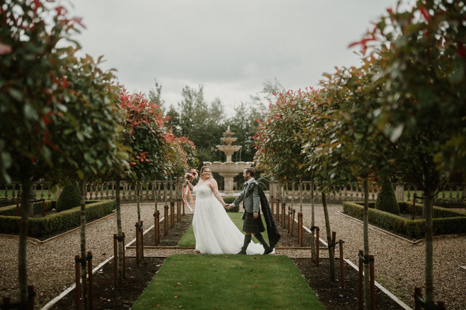 A wedding couple walks through a garden path lined with flowering trees.