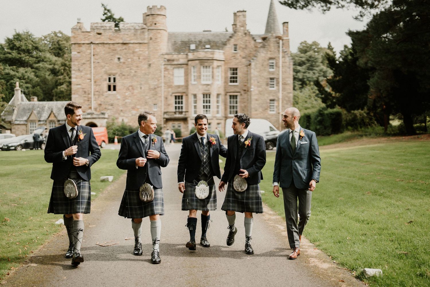 Groomsmen in traditional Scottish kilts walking outside a stone castle.