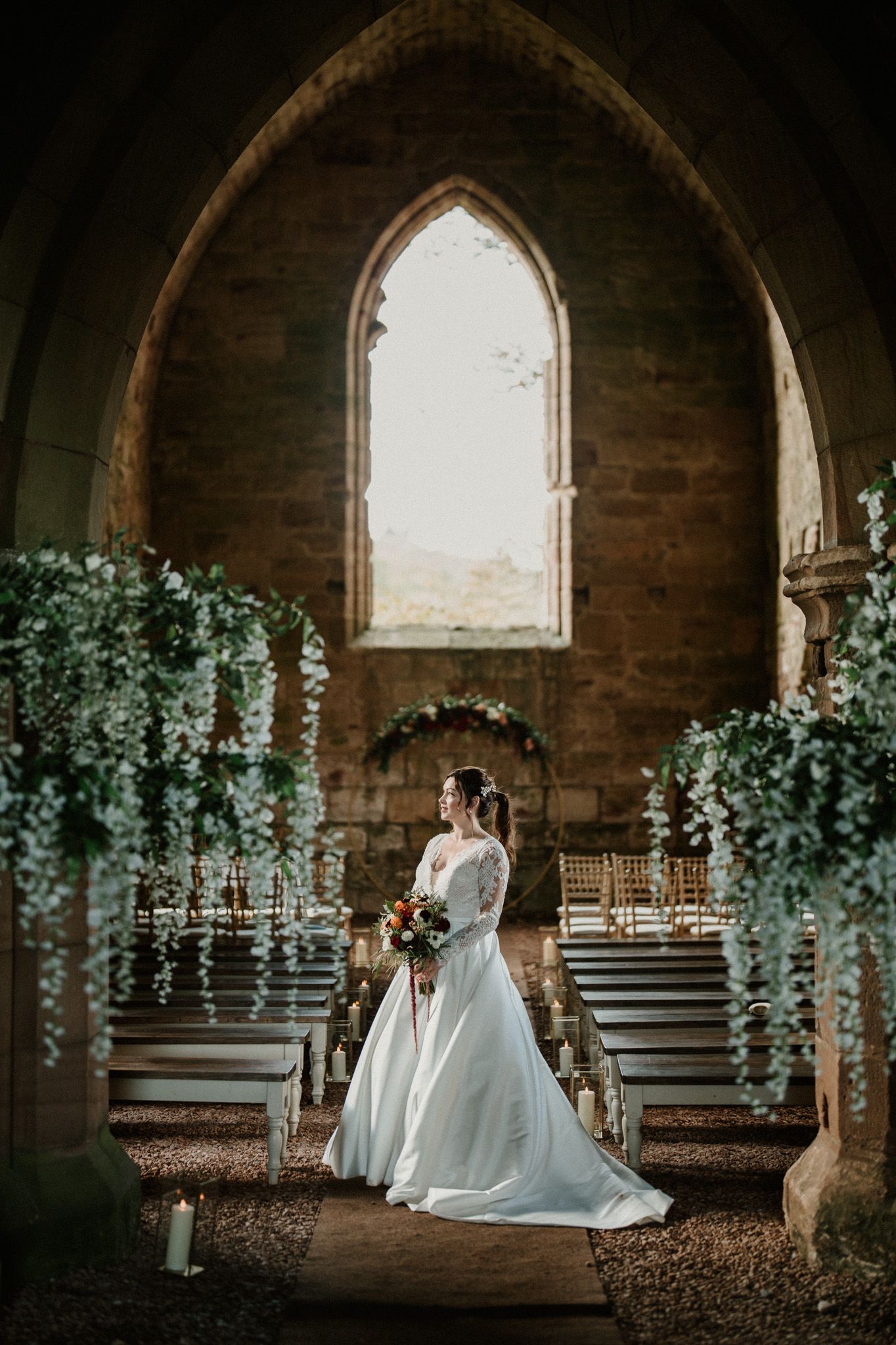 Wedding ceremony setup in gothic stone church with hanging greenery and wooden chairs.