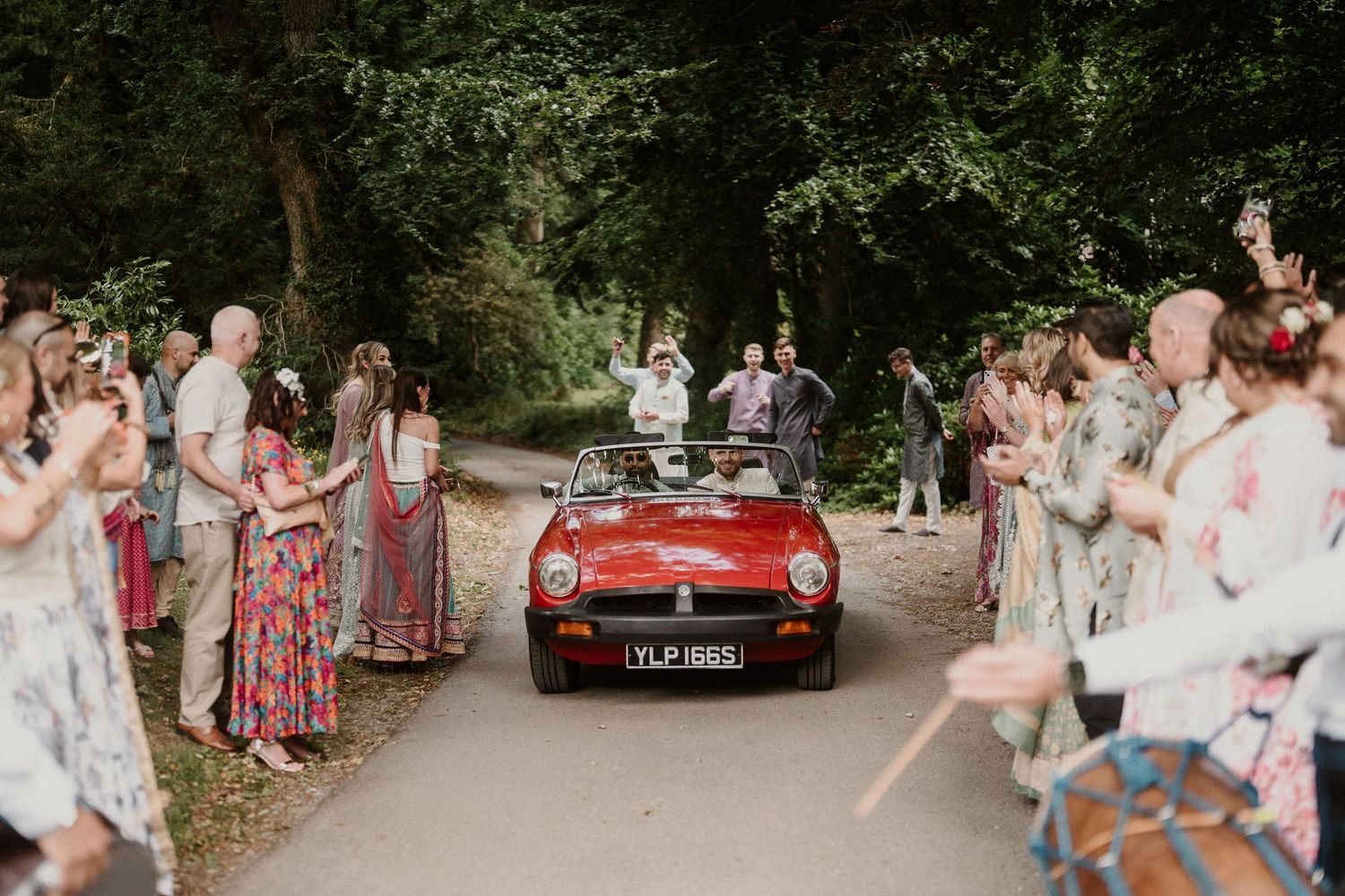 Wedding guests enjoying outdoor celebration with vintage red car in background.