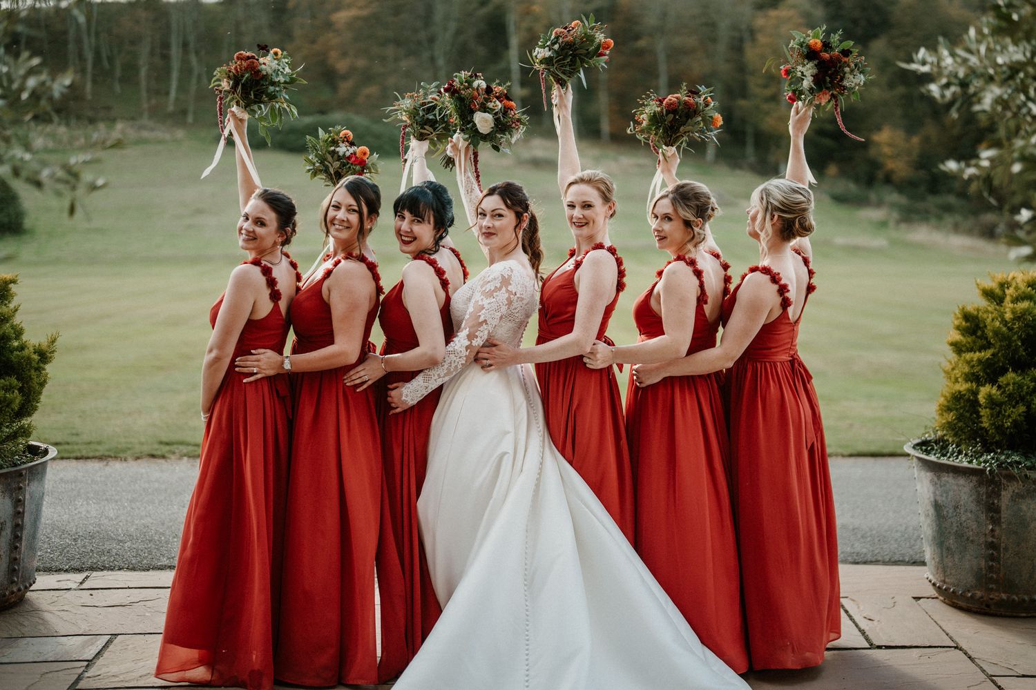 Wedding party in vibrant red bridesmaid dresses and white bridal gown pose together on stone patio.