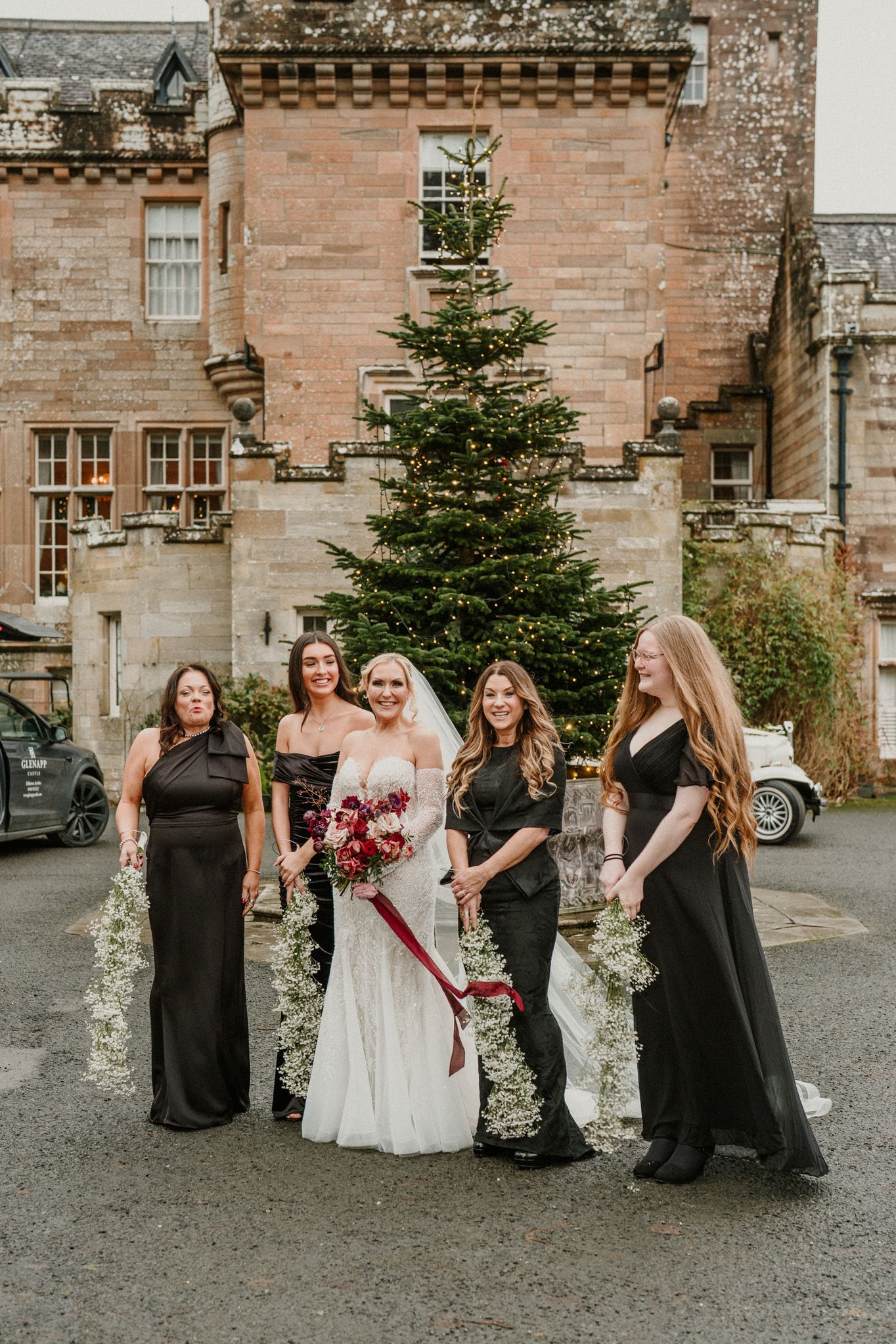 Wedding party posing outside a historic stone castle with evergreen trees.