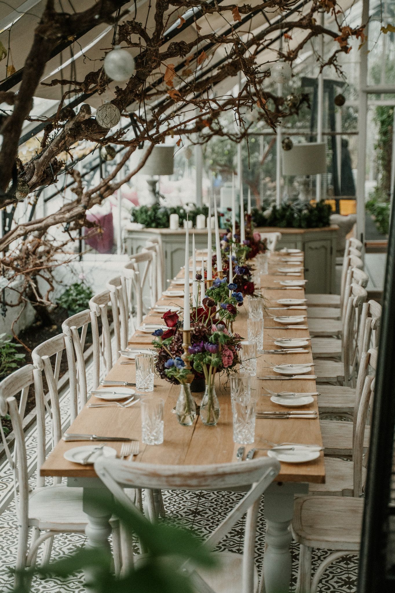 Long wooden dining tables adorned with floral arrangements and place settings in a sunlit greenhouse space.