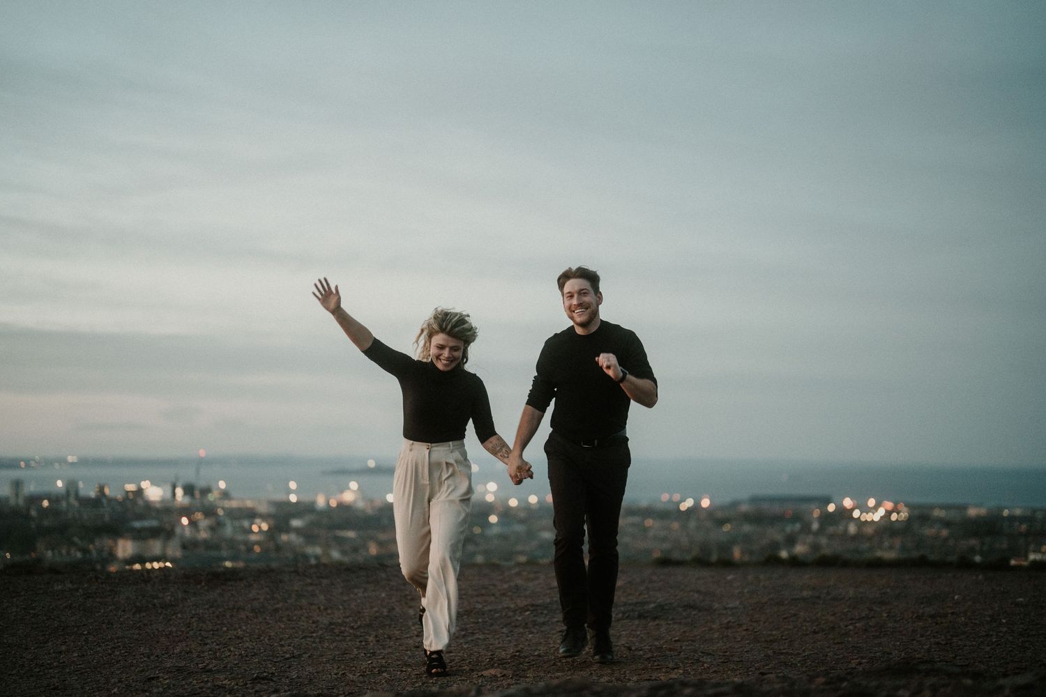 A joyful couple running hand in hand on a hilltop at twilight with a city lights backdrop.