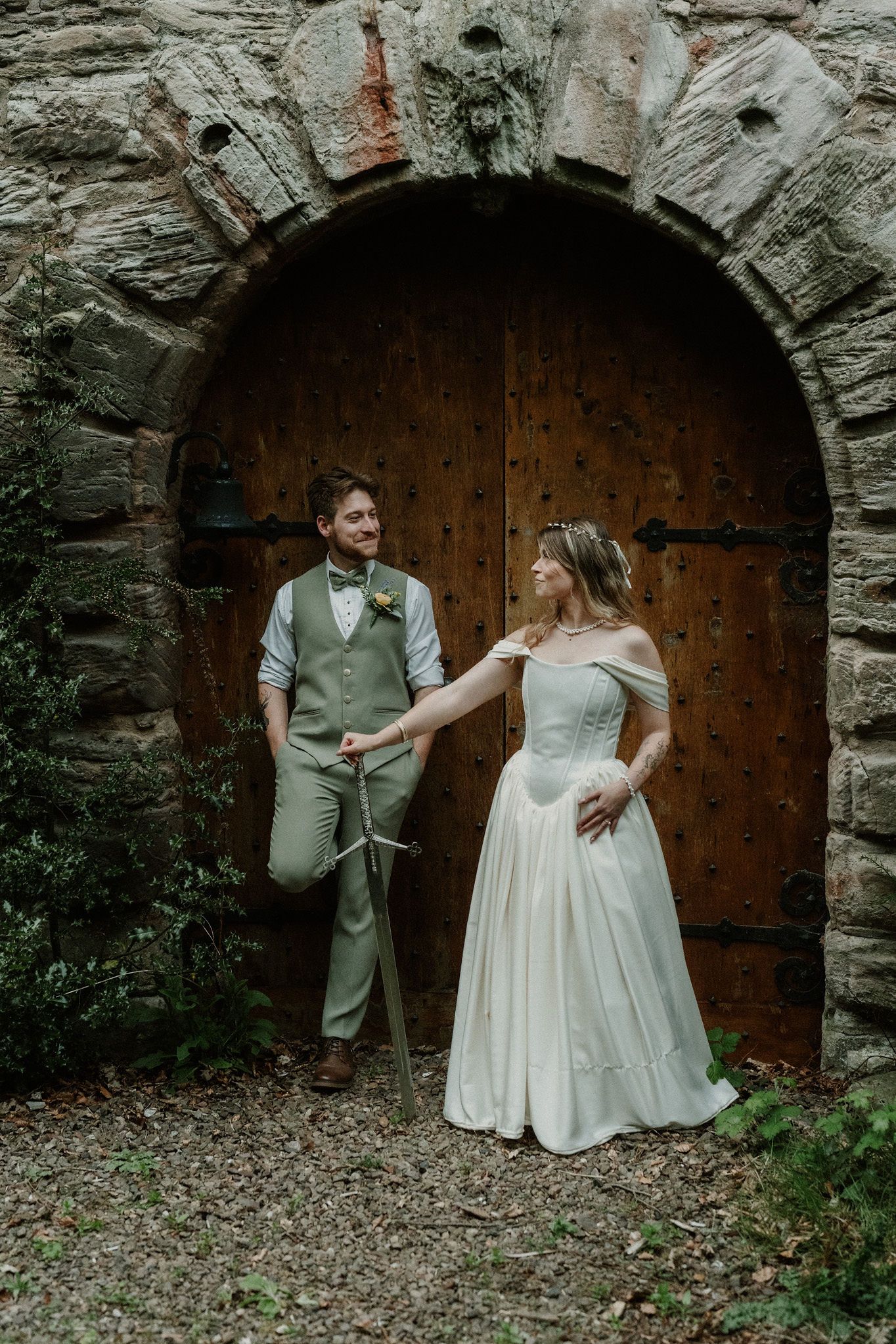 Bride and groom pose before a rustic stone arch with wooden door, groom holding a sword on their wedding day.