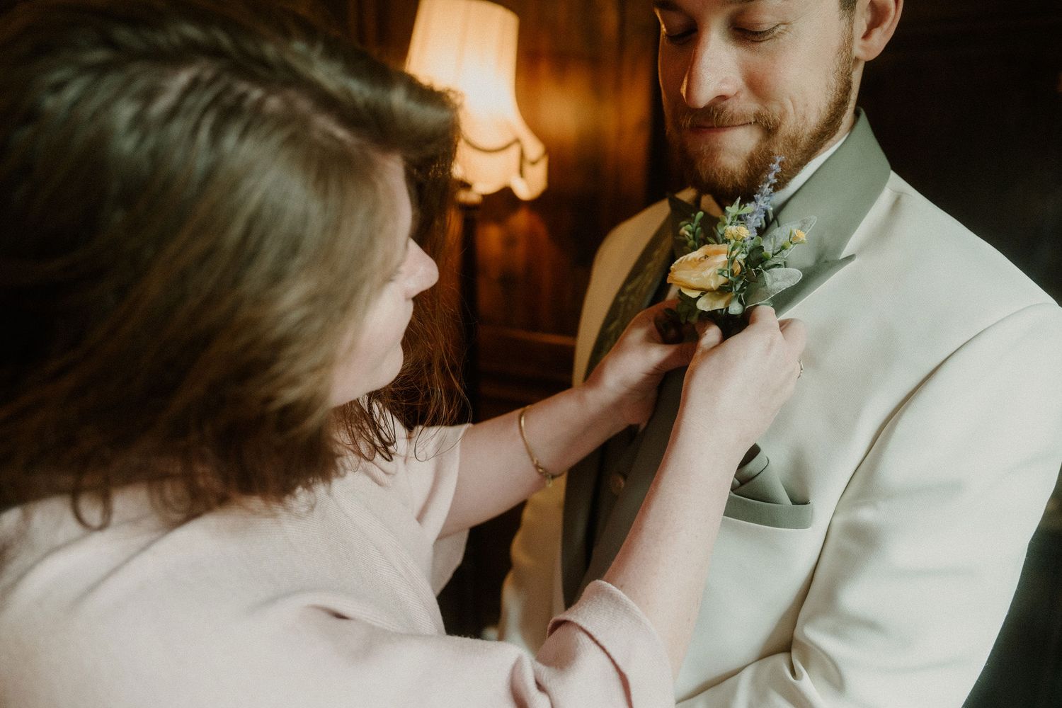 A woman pins a floral boutonniere on a bearded groom in a white suit before their wedding ceremony.