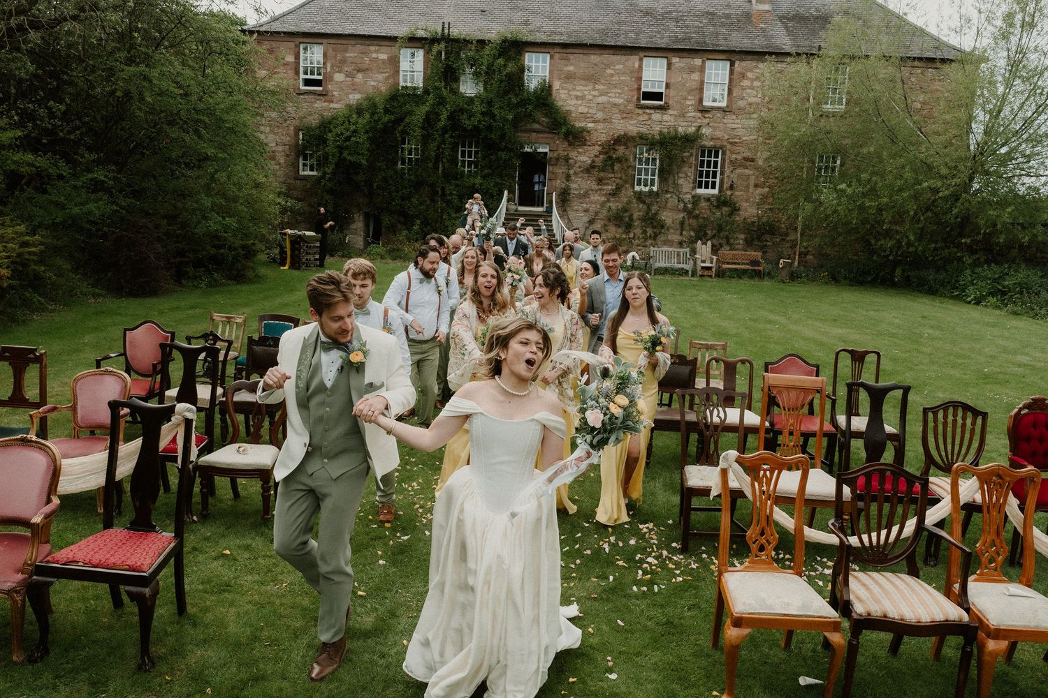 Joyful bride and groom lead wedding party across garden lawn outside historic stone manor house.