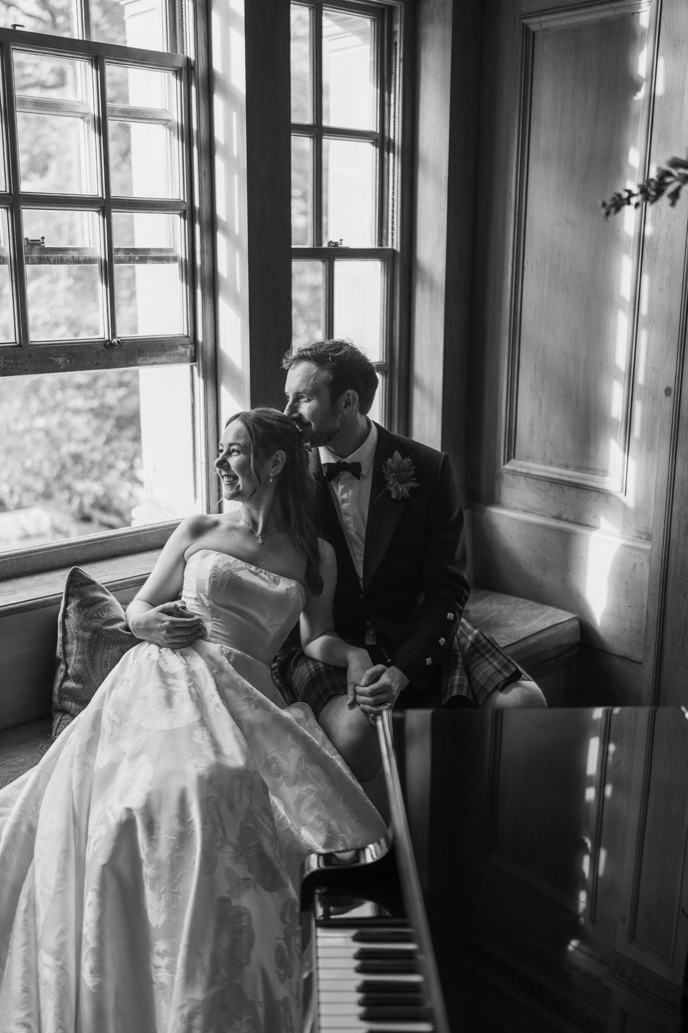 Black and white wedding photo of bride and groom sitting by a piano and large window, sharing a romantic moment.