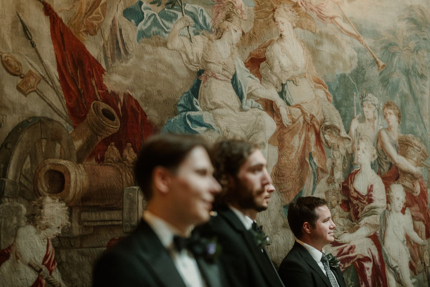 Three groomsmen in black tuxedos stand before an ornate baroque tapestry at an elegant wedding venue.