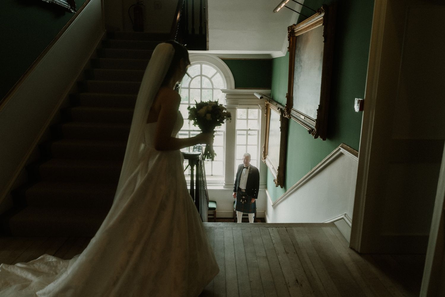 Bride in white gown and groom in kilt stand in elegant green-walled hallway with arched window and staircase.