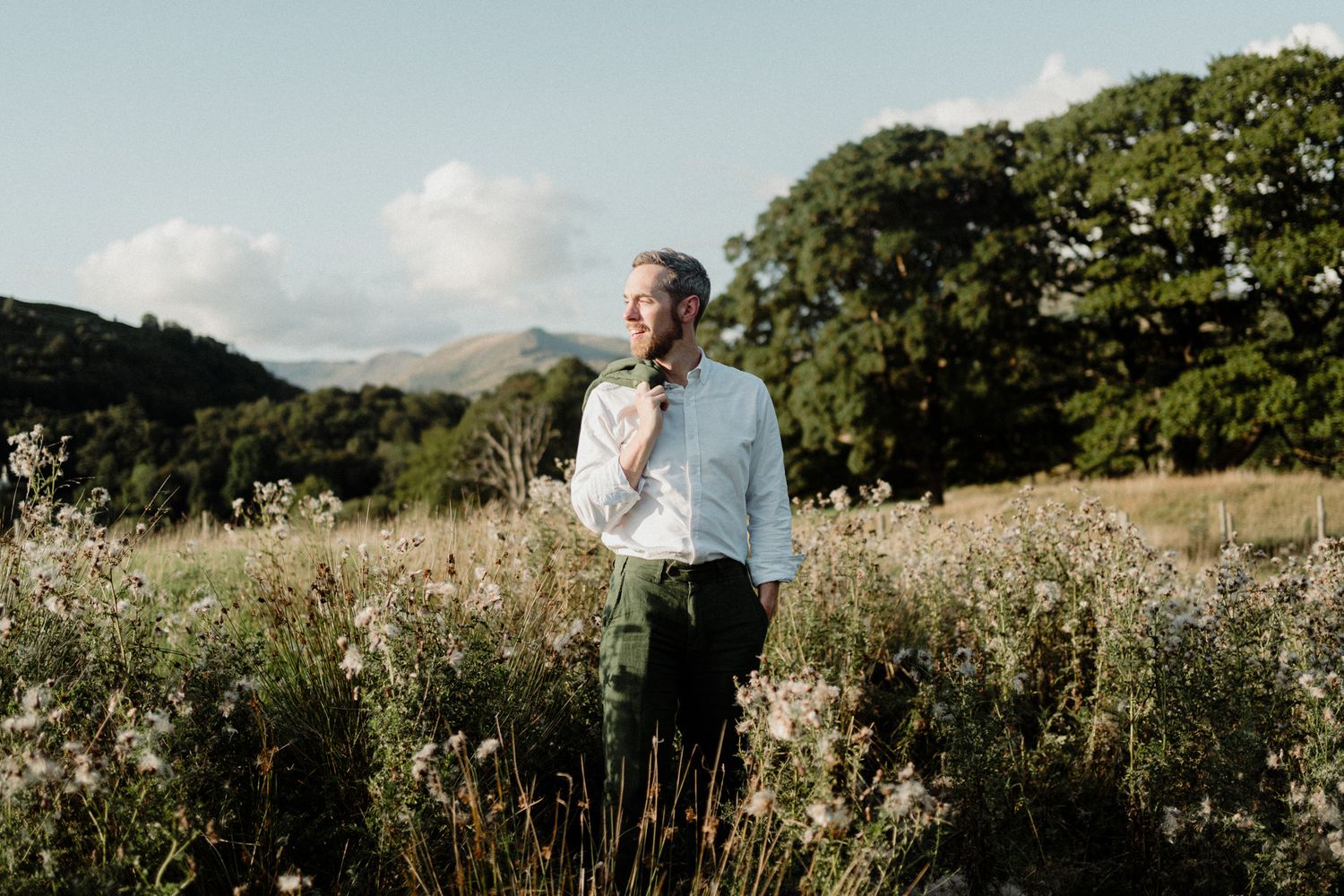 Man in white shirt standing in wildflower meadow with green hills and trees under blue sky.