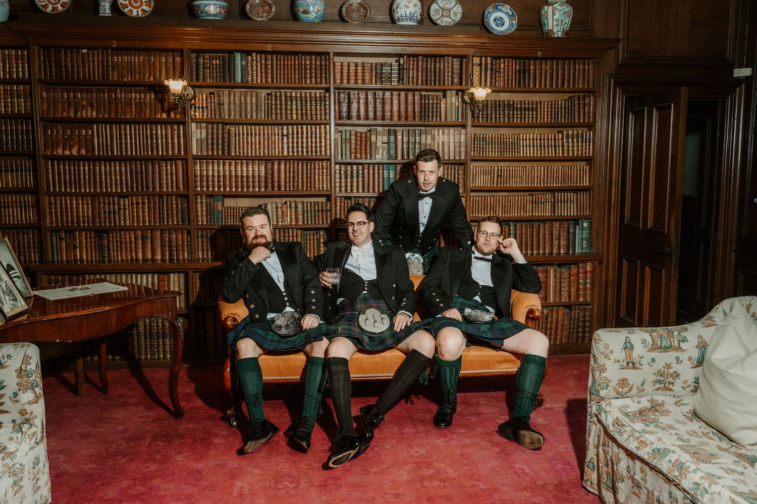 Four men in traditional Scottish kilts and green socks pose together in a classic wood-paneled library lined with antique books.