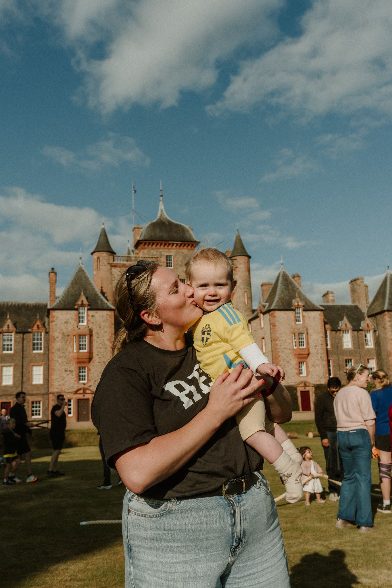 Woman in a black t-shirt kisses a smiling toddler in a yellow shirt on a lawn in front of a historic red brick castle under a blue sky.