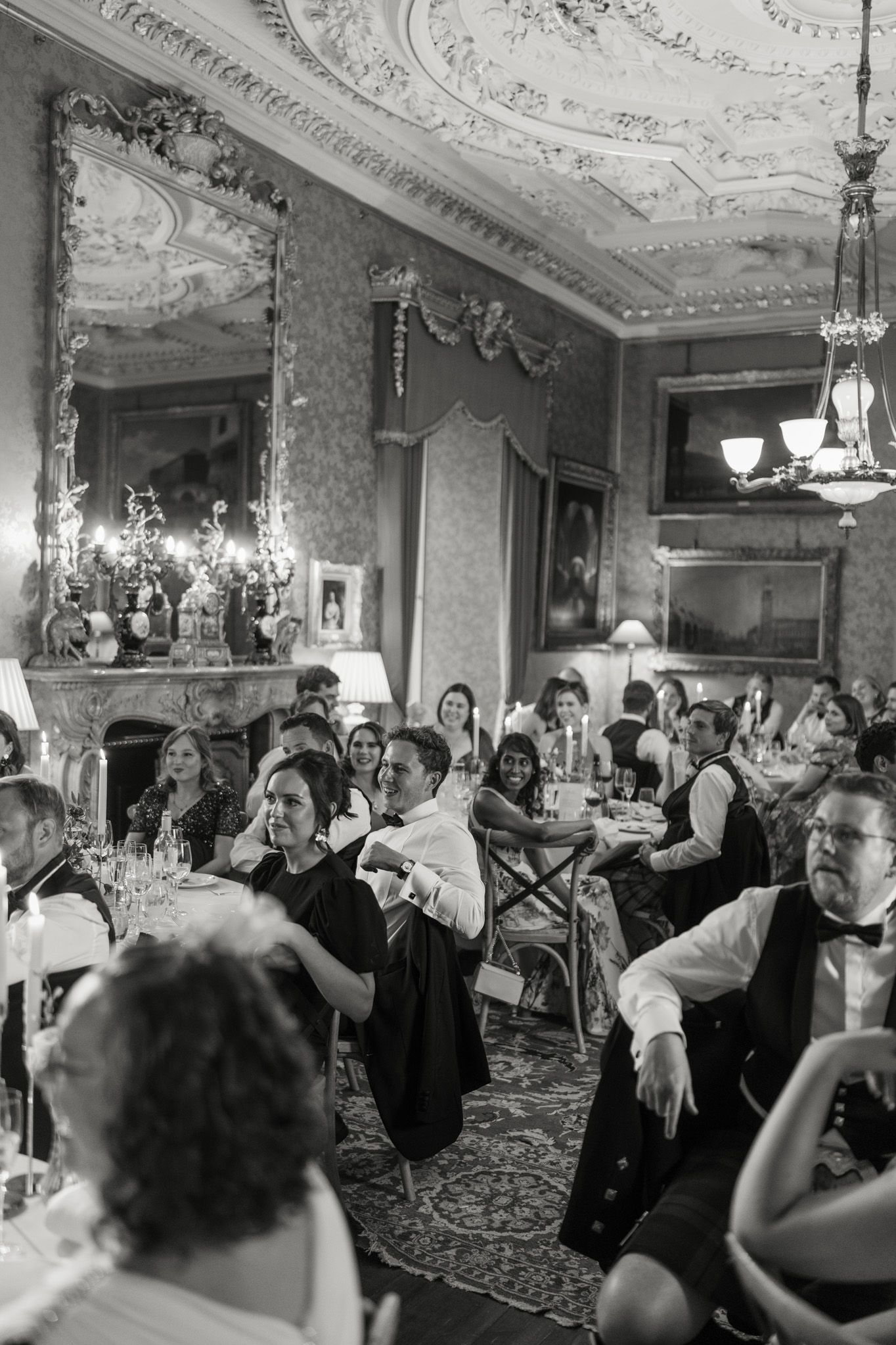 Black and white photo of wedding guests seated at dinner tables in an ornate room with a plaster ceiling, chandelier, and large mirror.