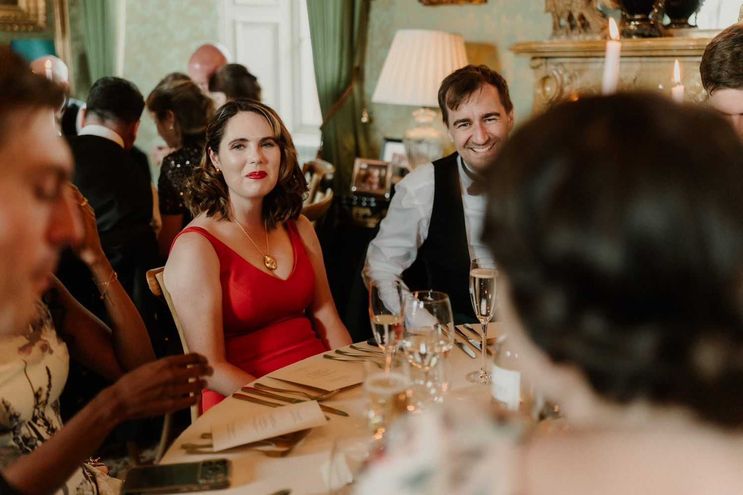 Smiling woman in a red dress and a man in a vest laugh at a candlelit reception table with wine glasses, surrounded by guests.