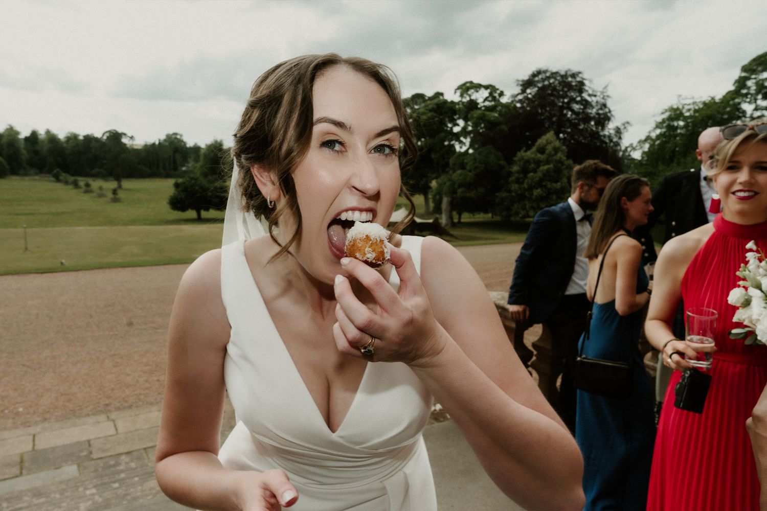 A bride in a white dress bites into a pastry at an outdoor wedding while guests chat behind her on a sunny lawn.