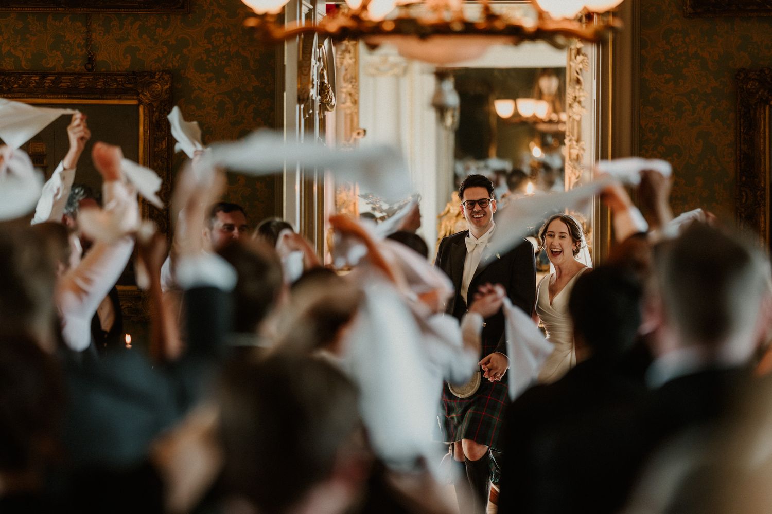 Wedding guests wave white napkins in a lively cheer as a bride and groom in a kilt walk through an ornate hall.