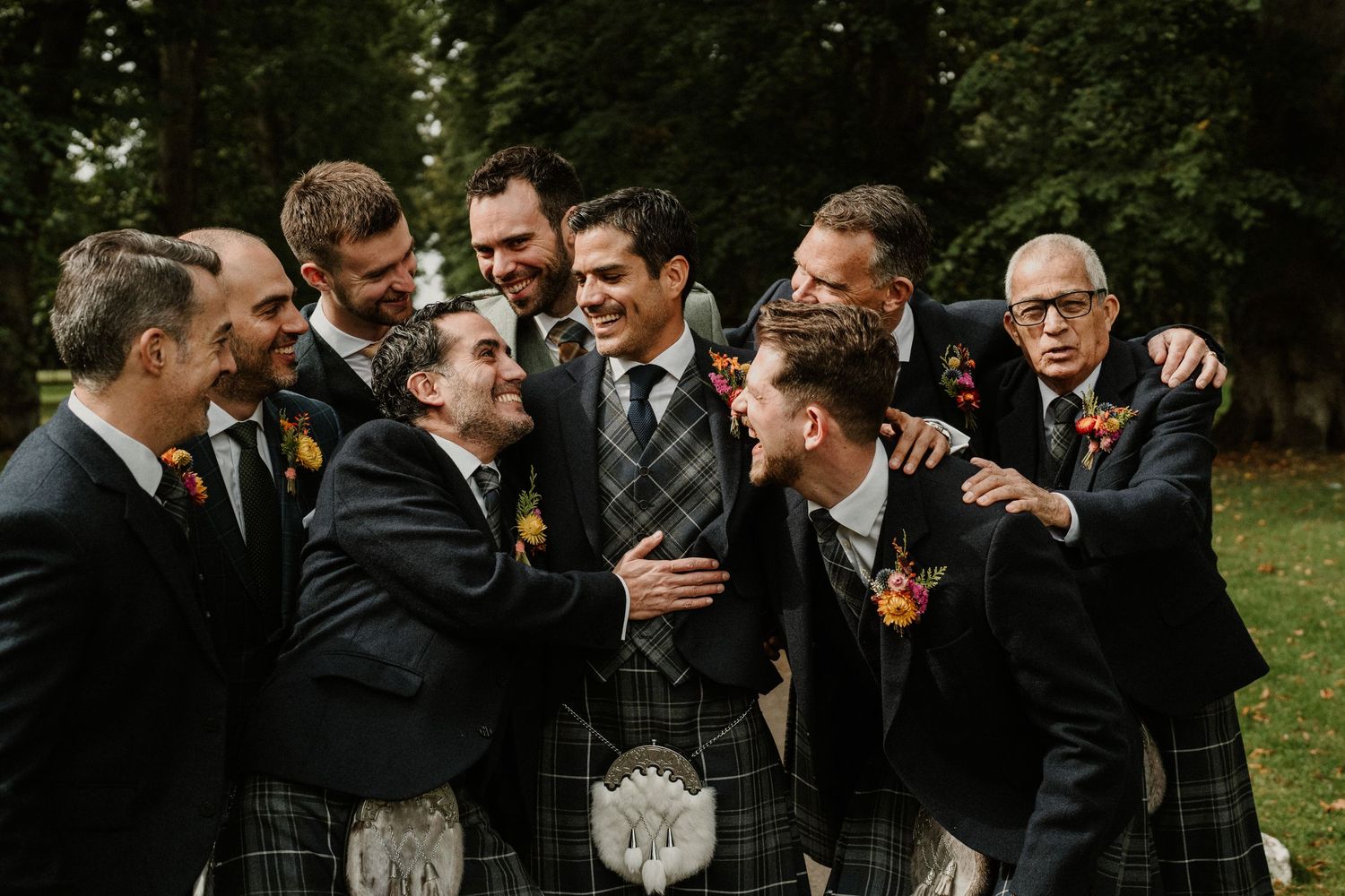 Groomsmen in traditional Scottish kilts and suits laugh together outdoors.