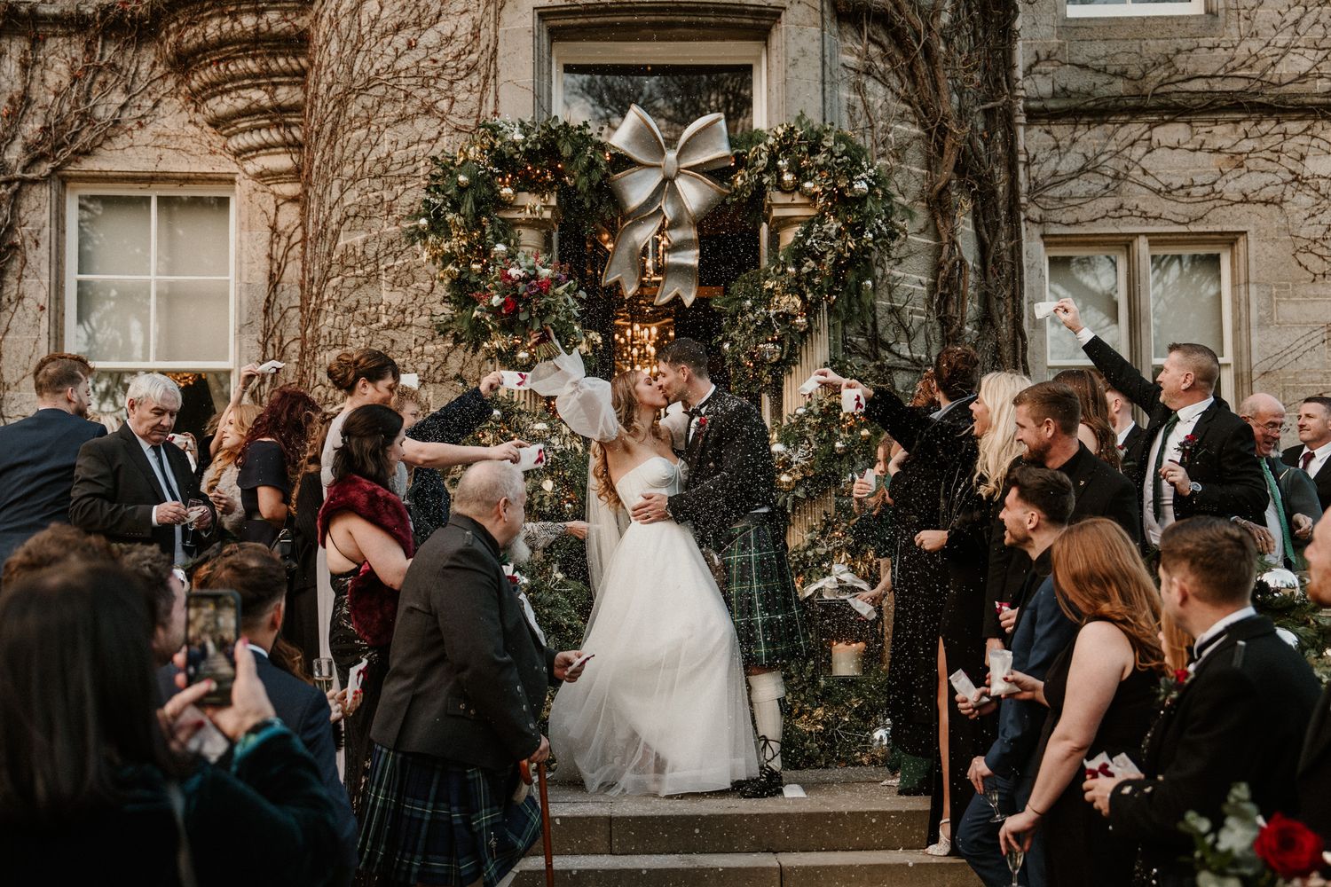 Wedding ceremony outside stone building with floral arch and gathered guests.