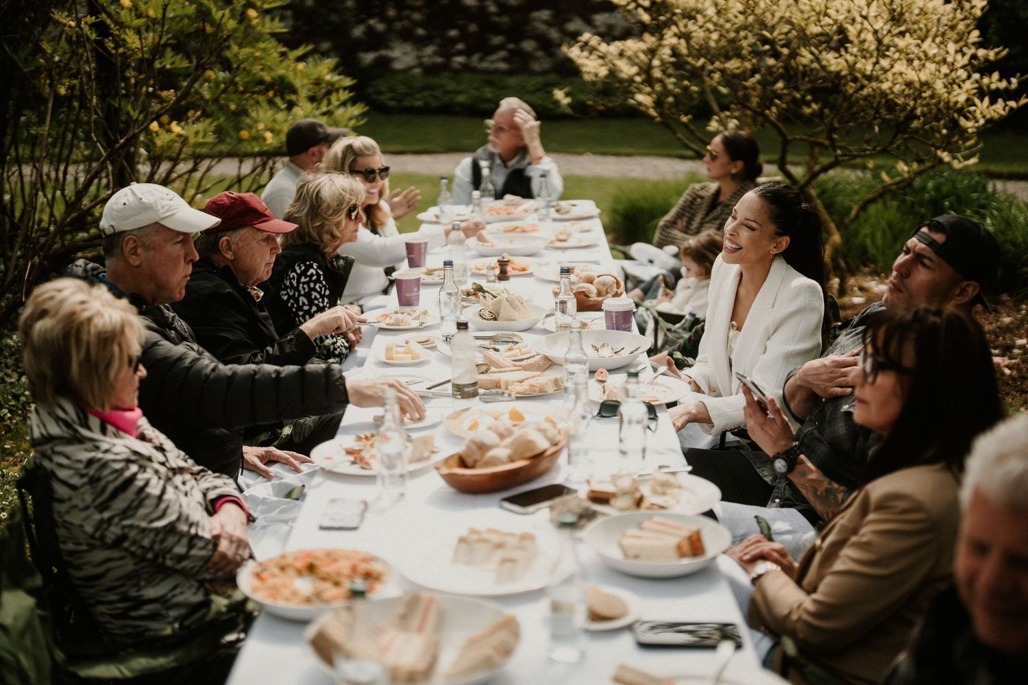 Candid outdoor dining scene with guests enjoying a meal at a long table in a garden setting.