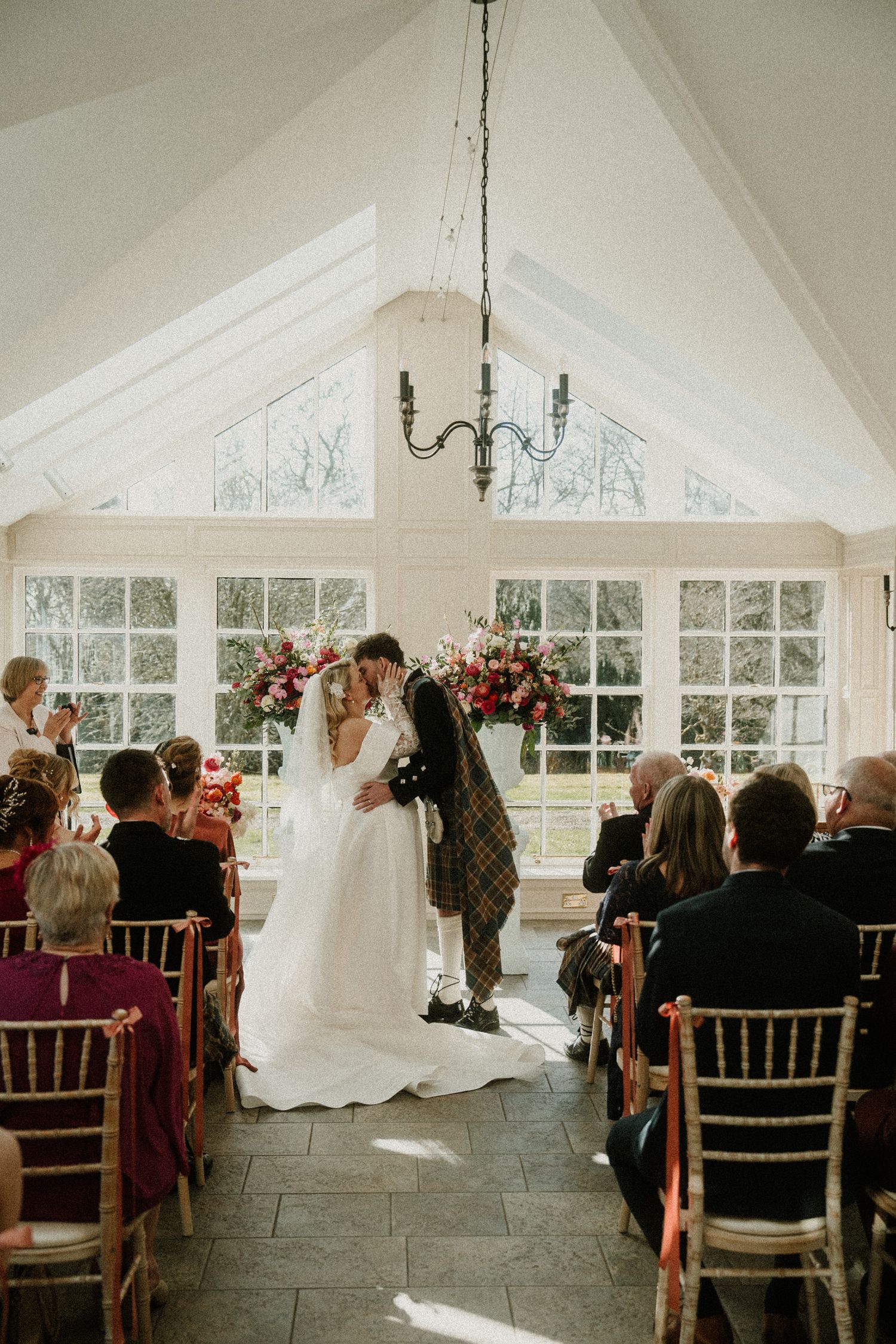 A Scottish wedding ceremony takes place in a bright conservatory with large windows and floral arrangements.