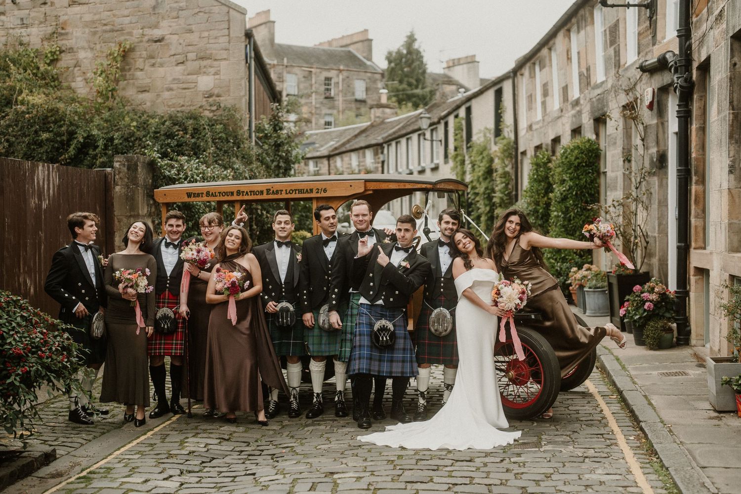 Wedding party gathered on a charming cobblestone street in traditional Scottish attire with kilts.