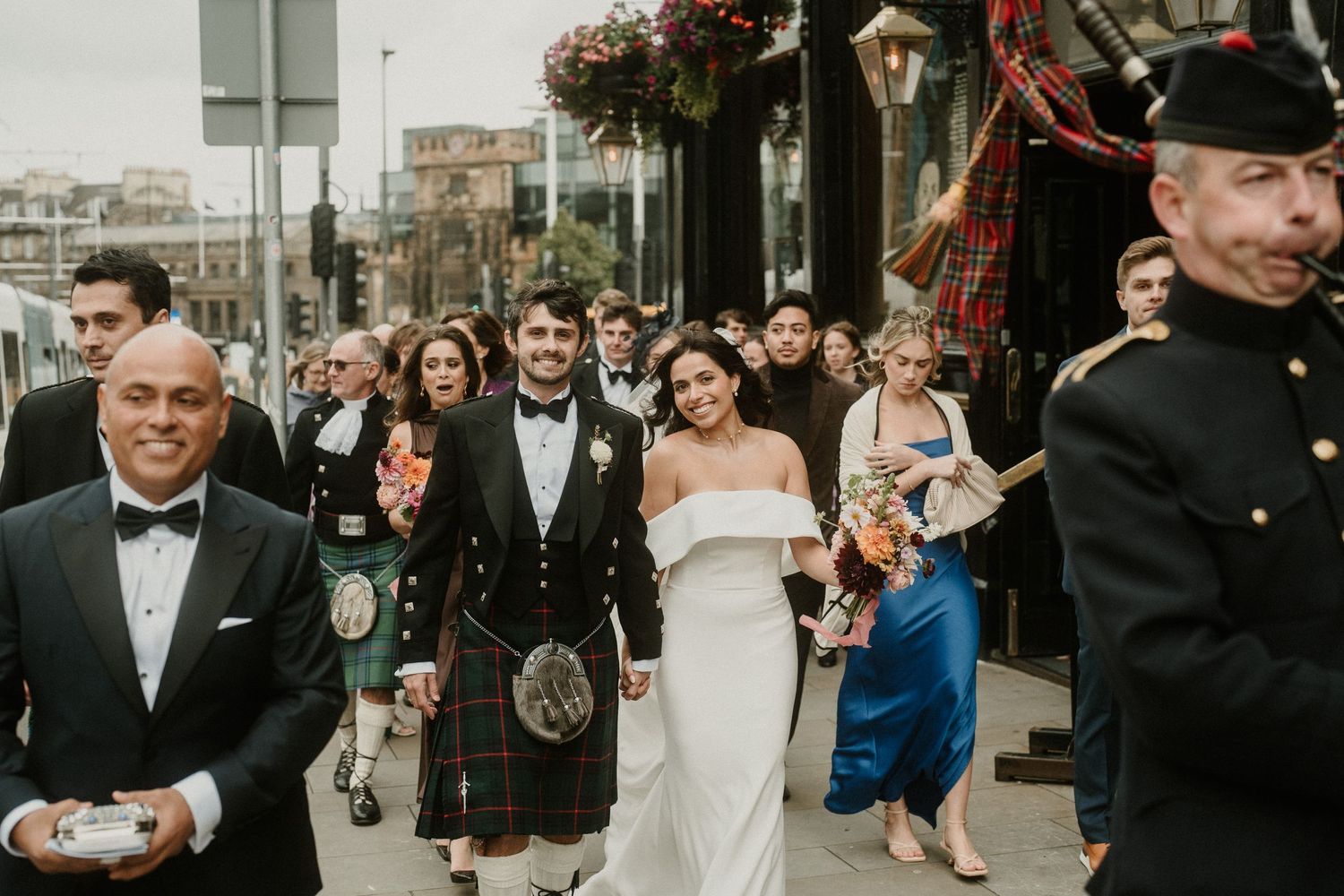 Outdoor wedding procession with bagpiper and guests walking along city sidewalk.